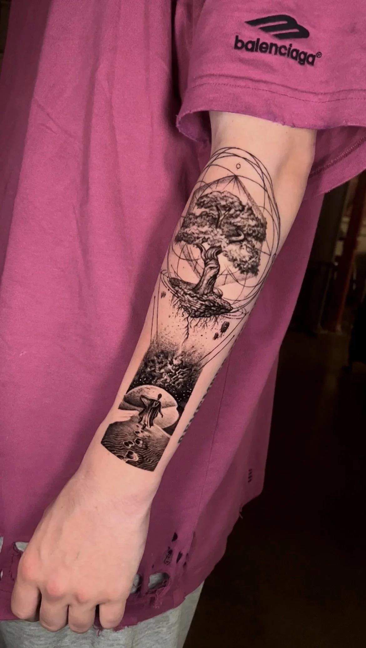 Black and gray tattoo on person's forearm depicting a tree with roots, a floating island, a person walking, and cosmic elements in a surreal style.