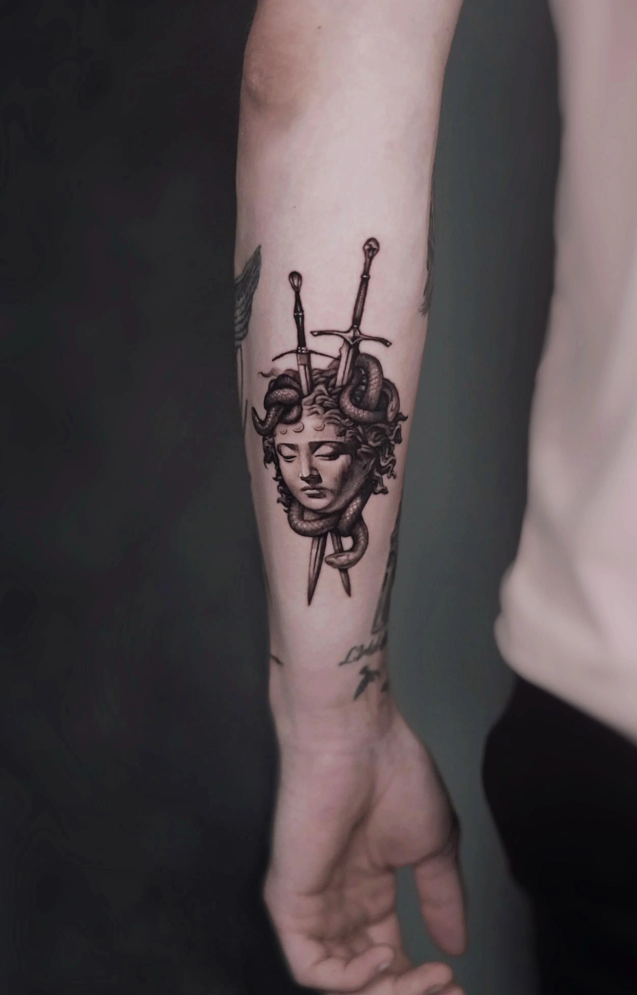 Tattoo of Medusa with snakes for hair, pierced by two swords, on person's forearm.