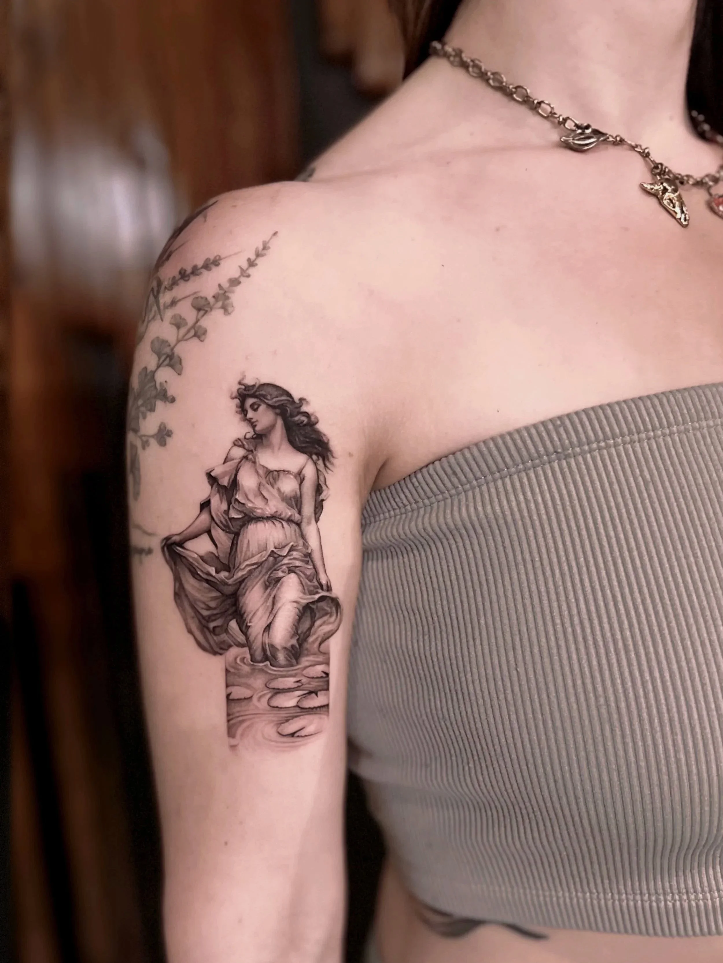 Tattoo of a woman in a flowing dress standing in water on upper arm, with leafy branch outline tattoo on shoulder, wearing a chain necklace, and dressed in a ribbed strapless top. Classical tattoo, realistic women tattoo, feminine fine line art.