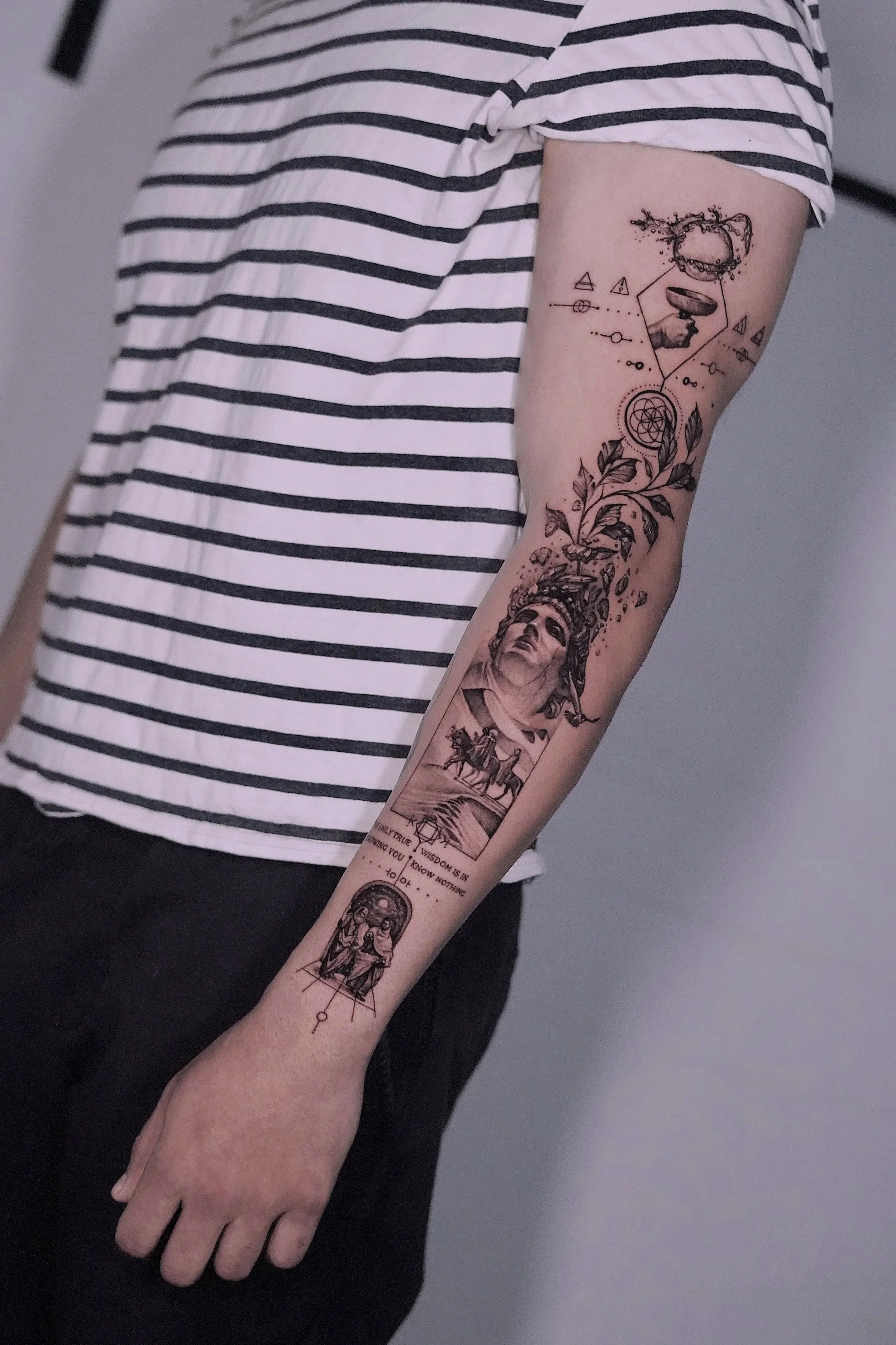 A person's arm with a detailed black ink tattoo sleeve featuring a classical statue, a horse, geometric shapes, an eye, and text, wearing a white and black striped short sleeve shirt. Geometric abstract tattoo, surrreal tattoo.