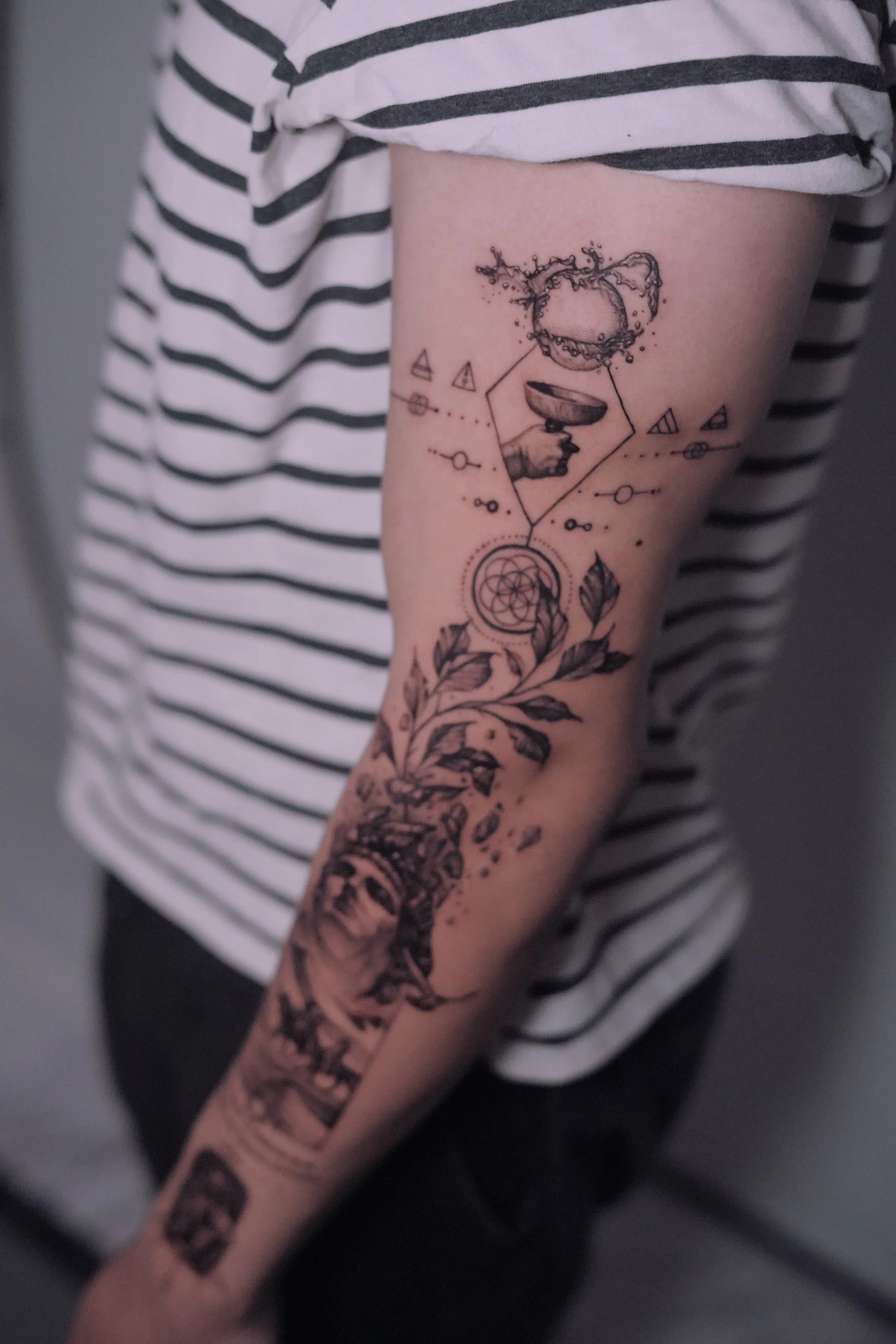 Black and gray tattoo on a person's arm featuring abstract geometric shapes, a hand holding a chalice with an orb or splash above it, a dreamcatcher, leafy plant elements, and a vintage woman's face at the wrist.
