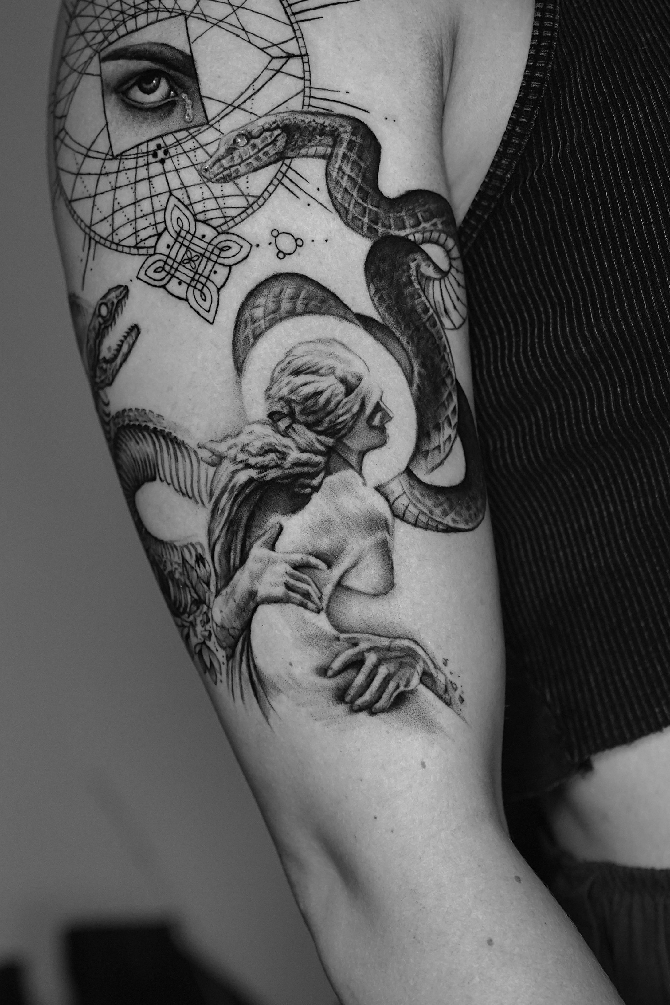 Black and white tattoo on a person's upper arm depicting a woman with long hair, a snake, a dreamcatcher with an eye and teardrop, a small crocodile, and an atom symbol, all in a detailed artistic style.