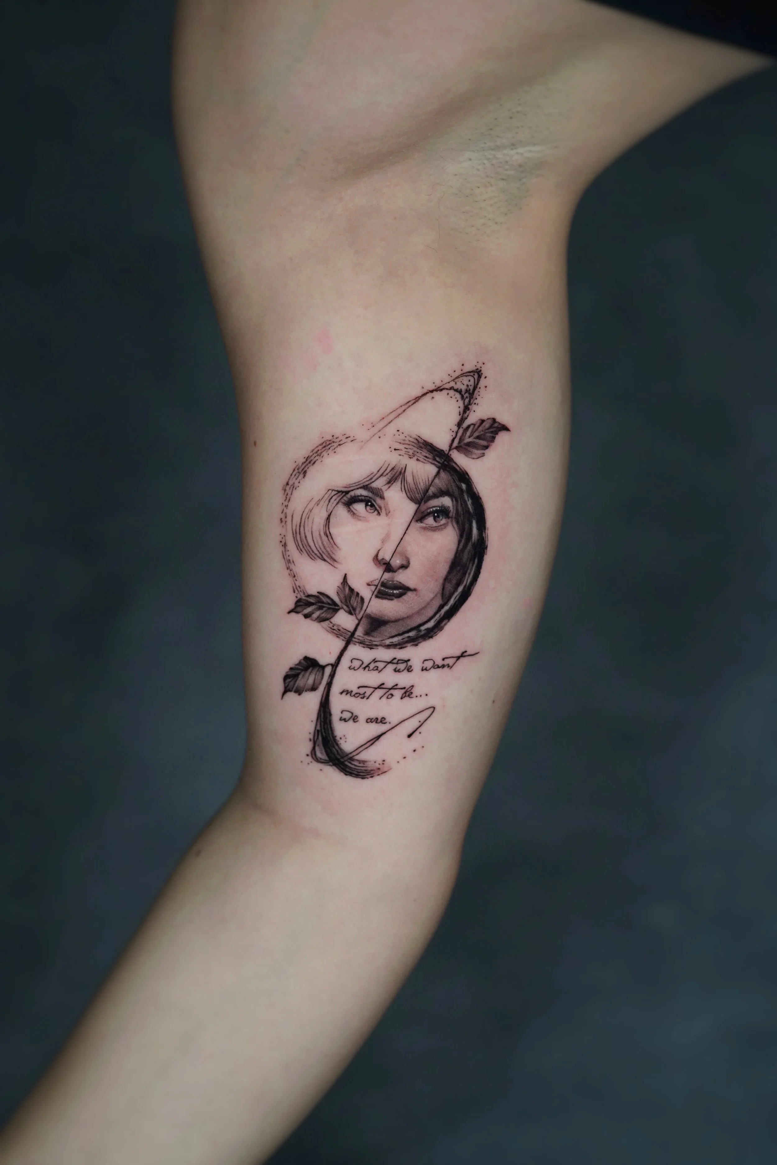 A tattoo on a person's arm featuring a woman's face surrounded by abstract lines, leaves, and a quote that reads, 'What do want most to be... We are.'