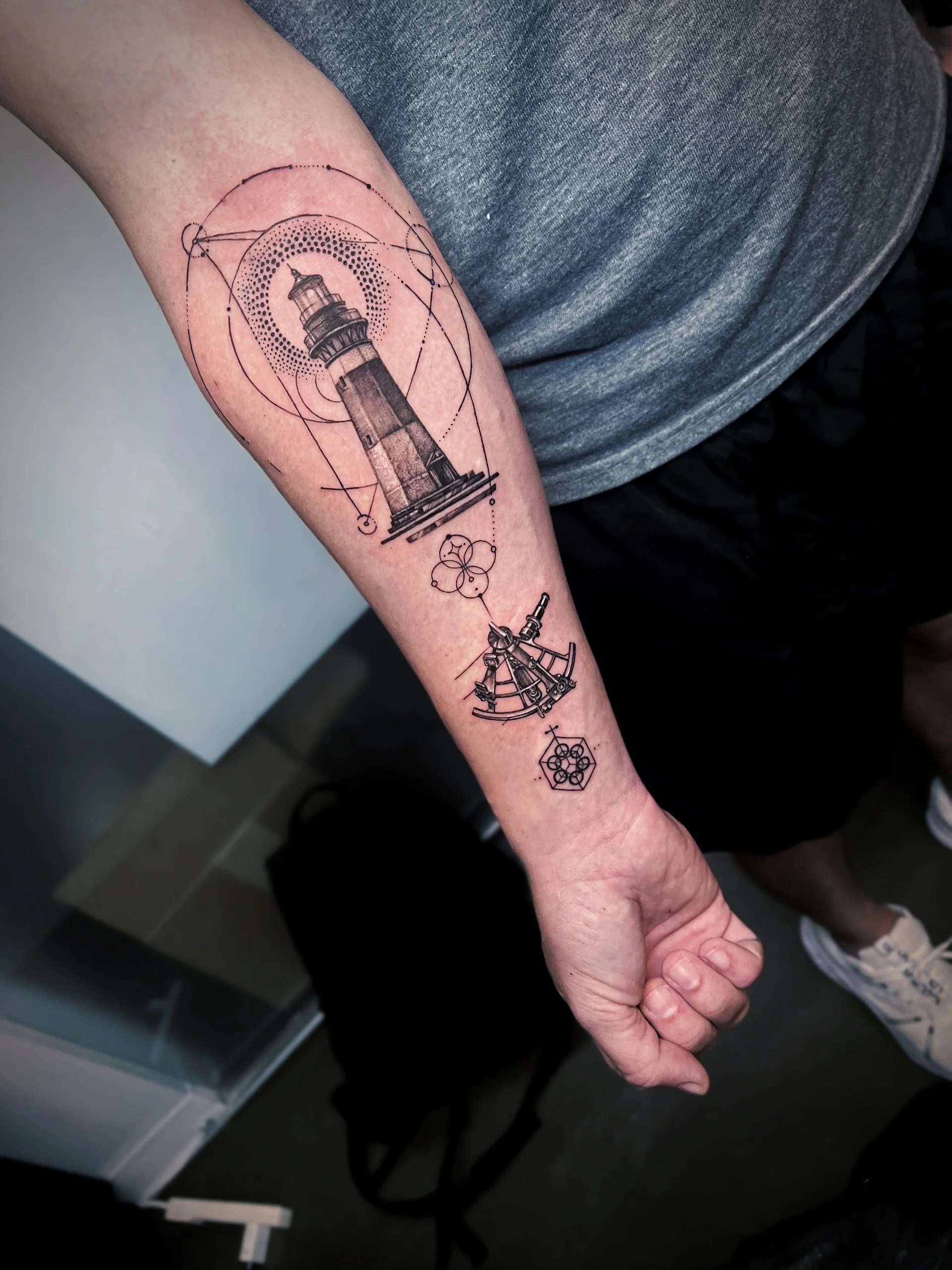 Tattoo of a lighthouse with geometric design elements on a person's forearm.