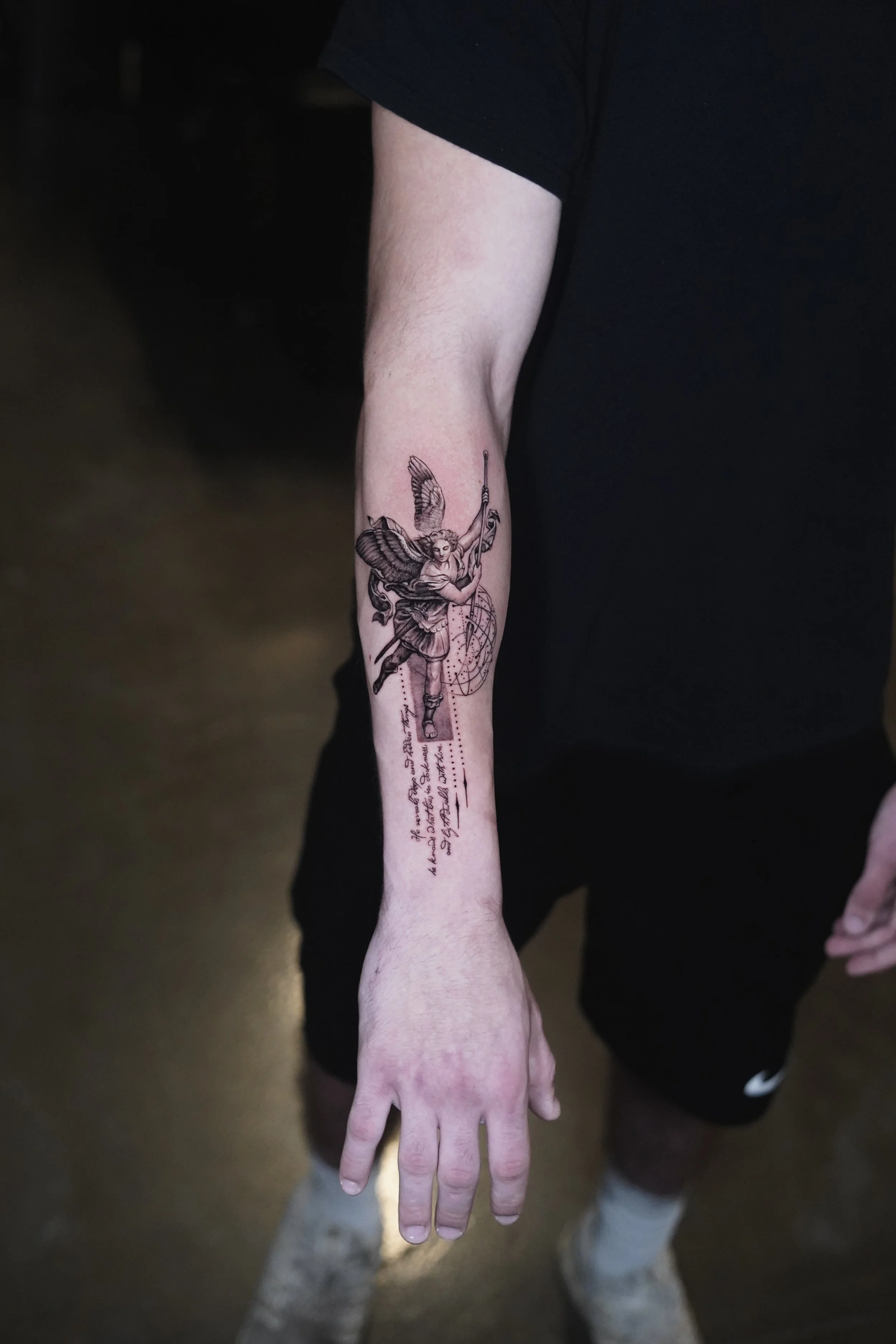 A person's forearm with a detailed black and gray tattoo of a winged woman holding a spear, standing on a geometric shape, with handwritten script and geometric designs below.