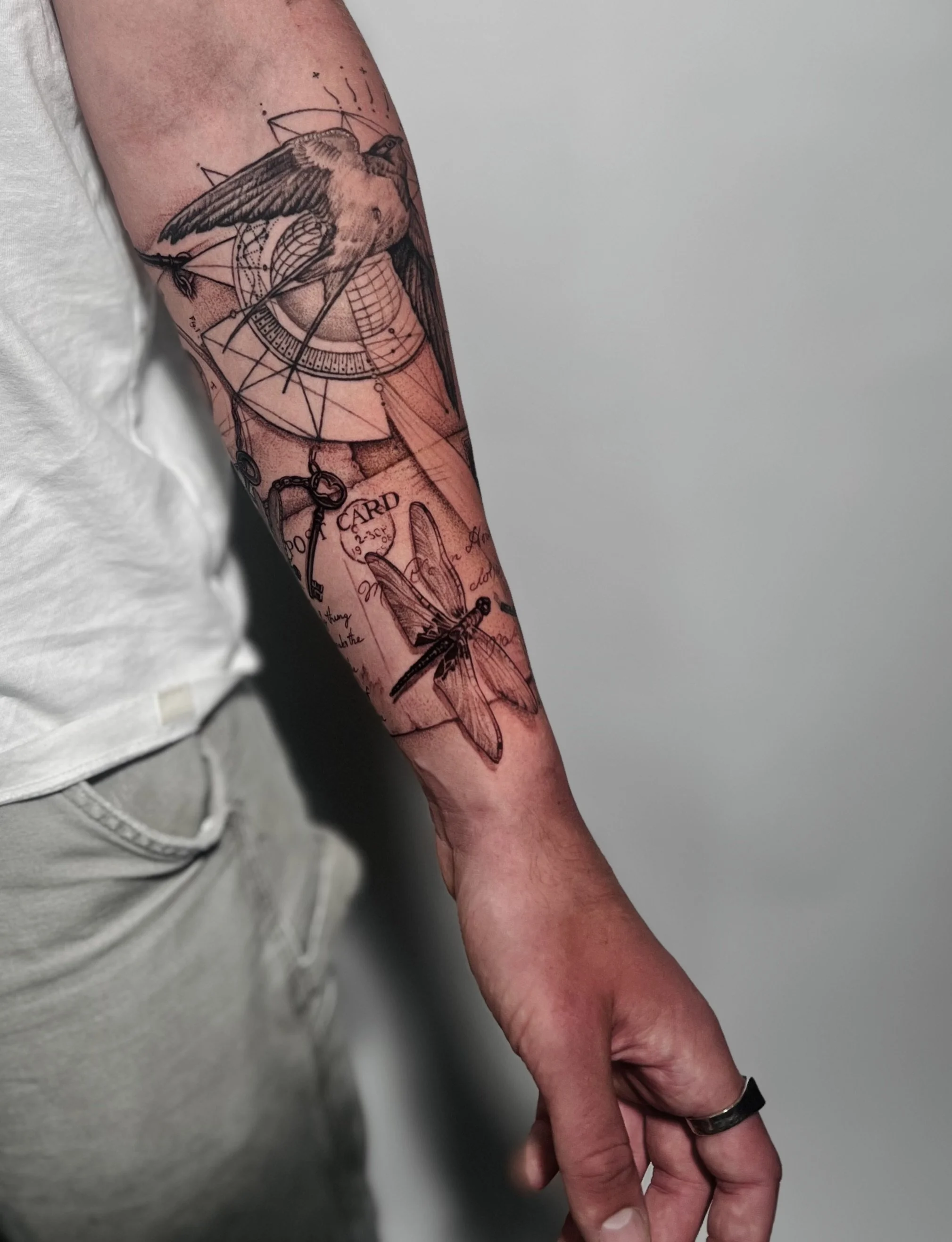 Tattoo of a bird and a dragonfly on a person's arm, with geometric and artistic designs, including a compass and handwritten text.