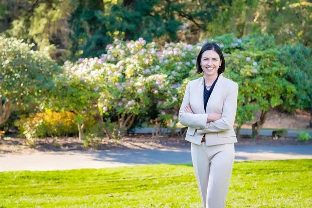 Judge Adrian Brown in a beige suit standing outdoors on grass with pink flowering bushes and trees in the background, smiling with arms crossed.