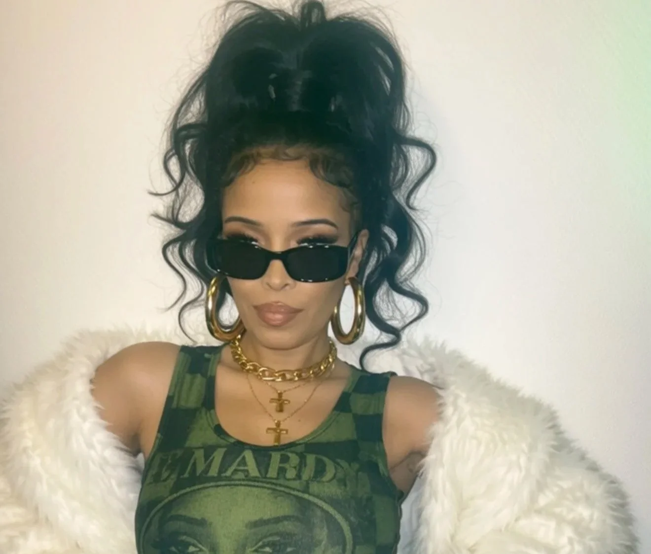 Pilar Scratch with dark curly hair styled in an updo, wearing sunglasses, large gold hoop earrings, layered gold necklaces, a sleeveless green graphic tank top, and a white fluffy jacket over her shoulders.