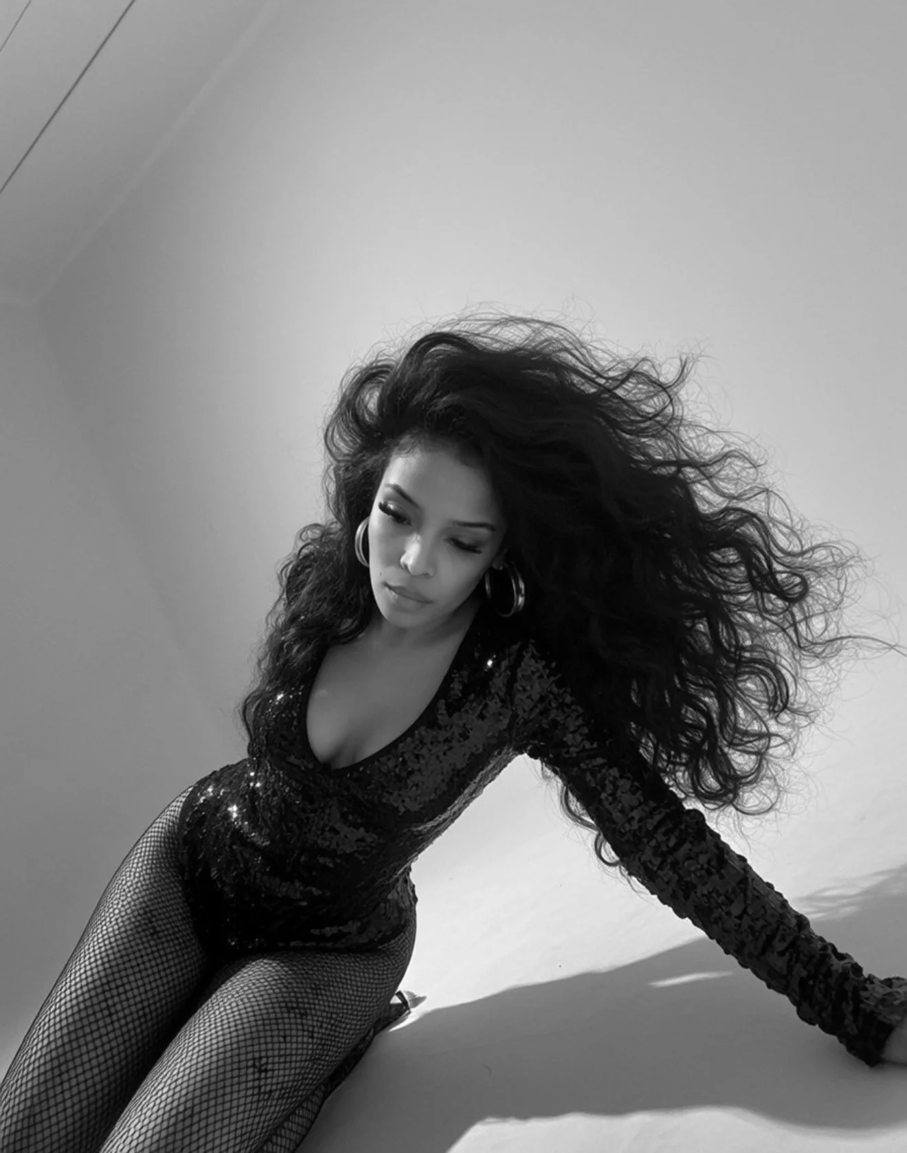Pilar Scratch with long, curly hair posing in a black sequin outfit and fishnet stockings, looking down, in a black-and-white photo.