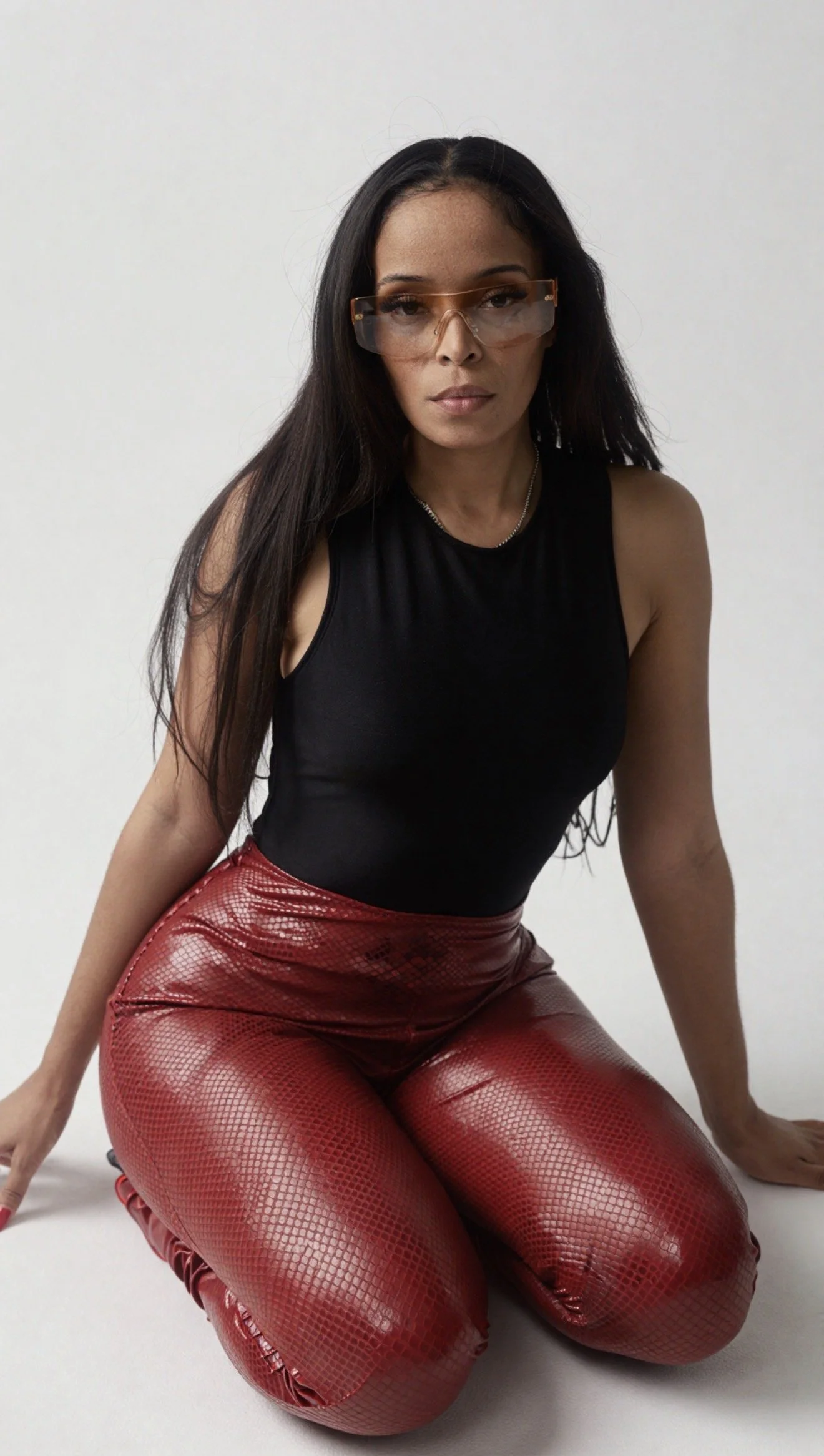 Pilar Scratch with long dark hair wearing oversized sunglasses, a black sleeveless top, and shiny red pants with a snakeskin pattern, kneeling on the floor against a plain white background.