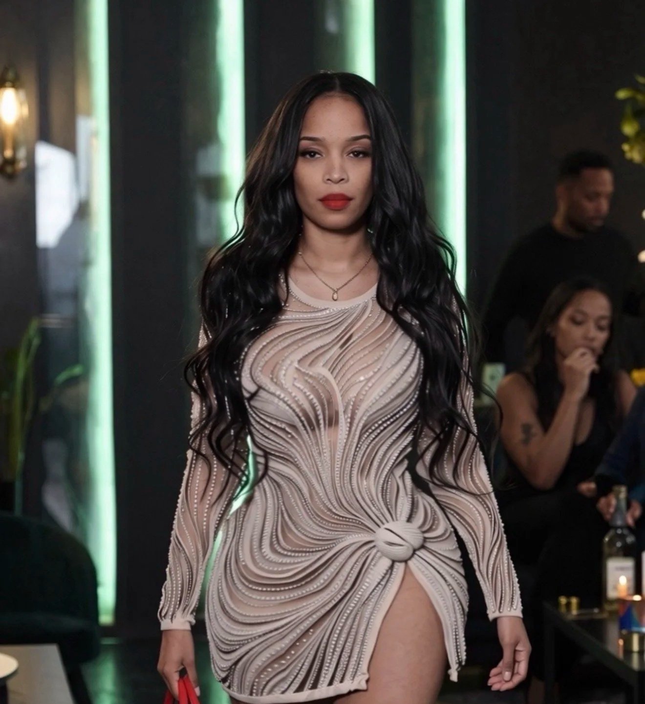 Pilar Scratch with long black wavy hair and red lipstick, wearing a nude-colored sheer dress with intricate beadwork and a slit, walking in a dimly lit indoor setting with other people in the background.