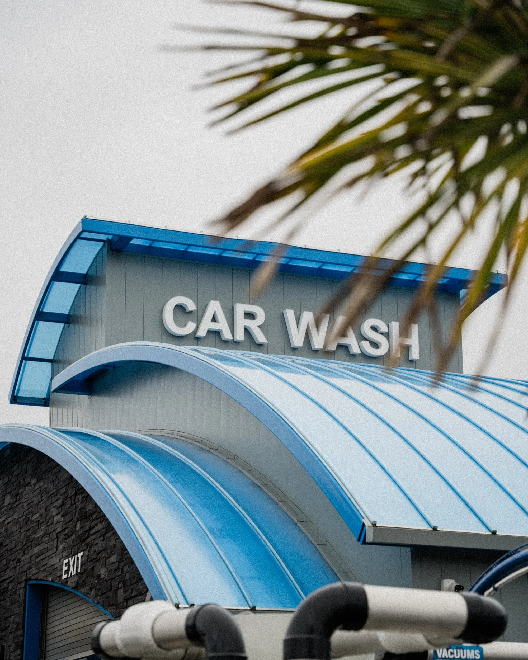 Building with a sign that says 'CAR WASH' and an entrance with a blue awning, with an exit door below it and a sign that says 'Vacuums' nearby, seen through neighboring objects and the edge of a parking lot in the pacific northwest.