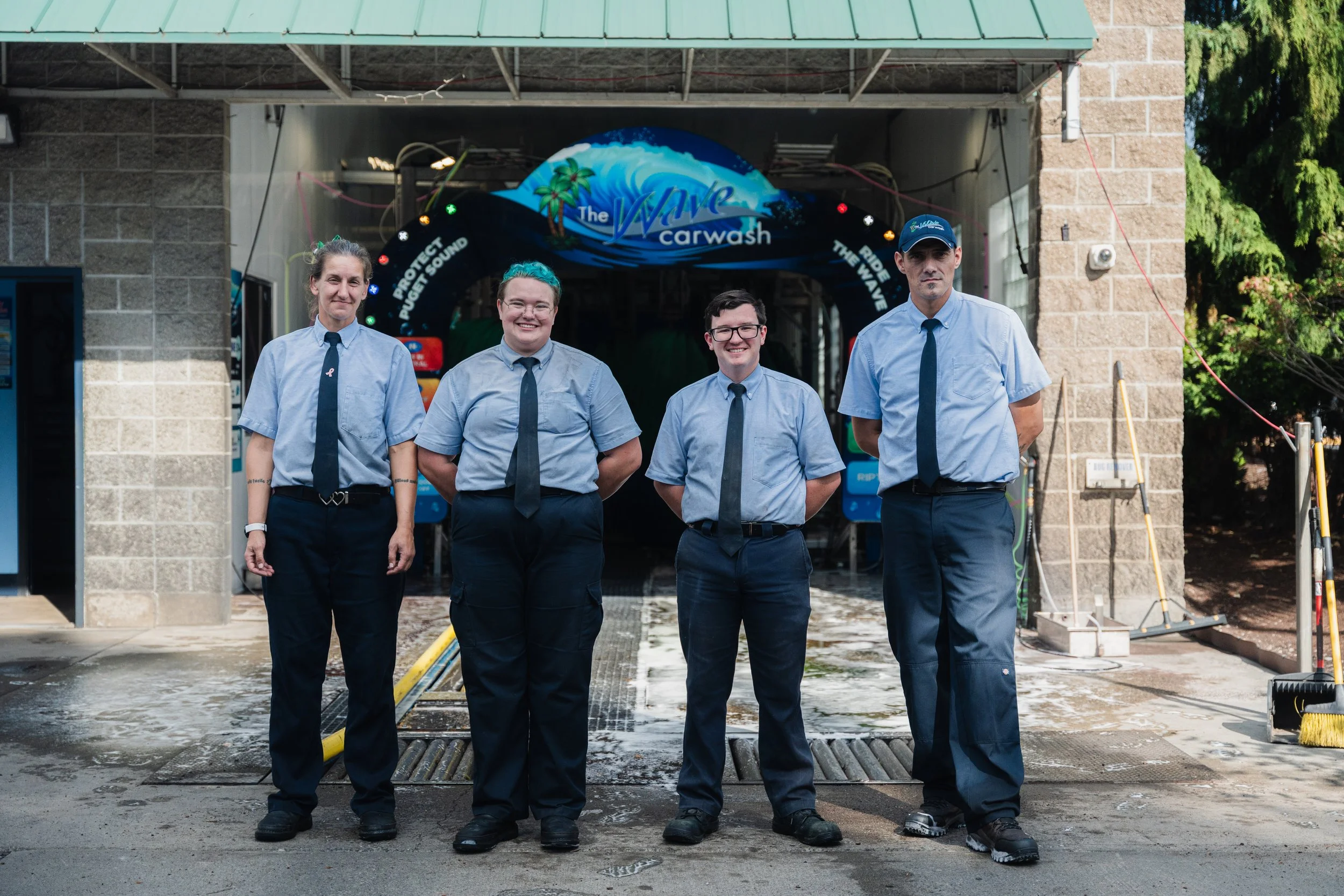 Four people standing in front of a car wash entrance, wearing uniforms with light blue shirts and dark trousers, with signs reading "Protect Quiet Sound" and "Ride The Wave" in the background with the Pacific Northwest.