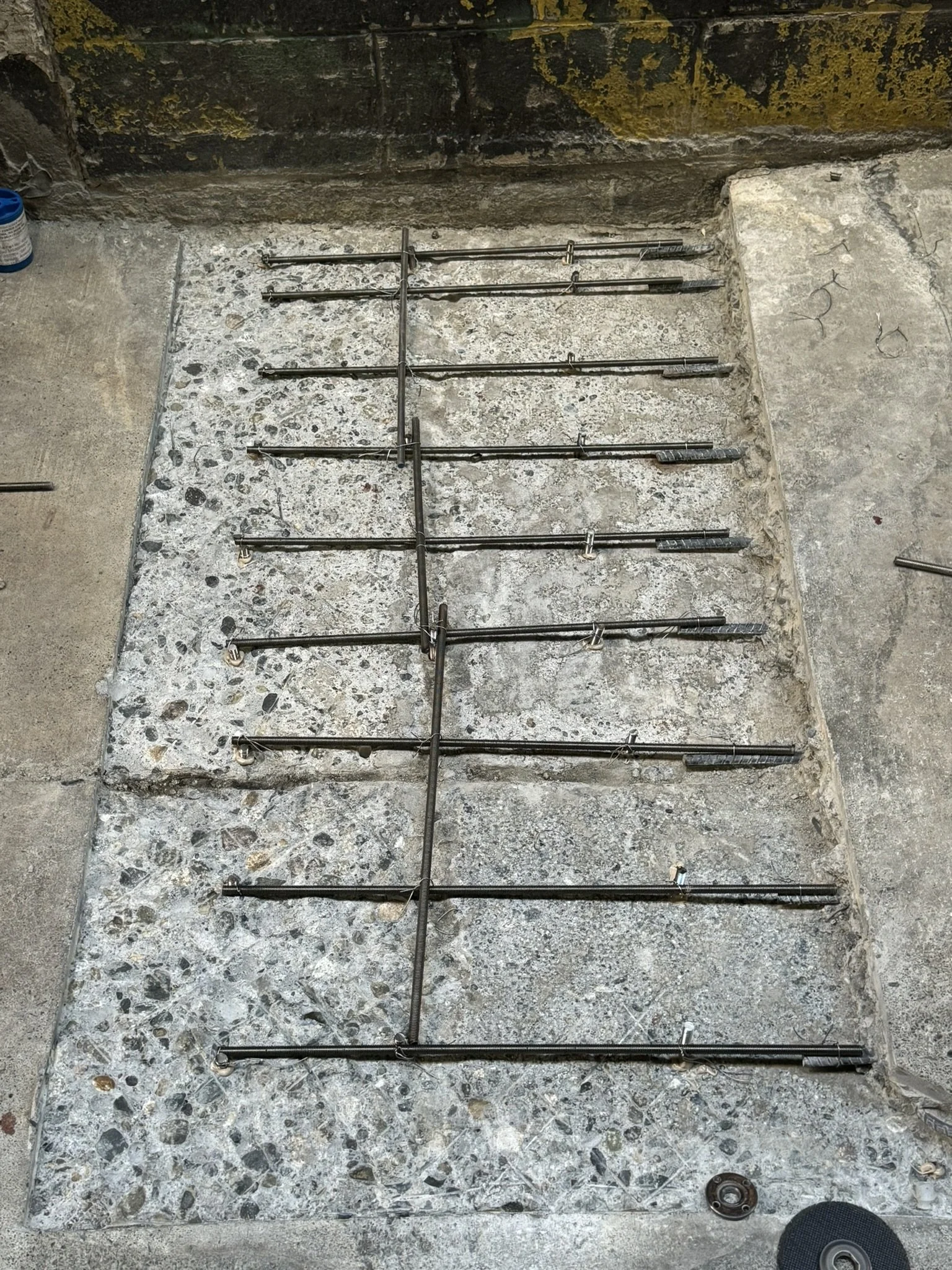 Metal rebar arranged on a concrete floor for construction, with a pointed-tapered object at the bottom right corner.