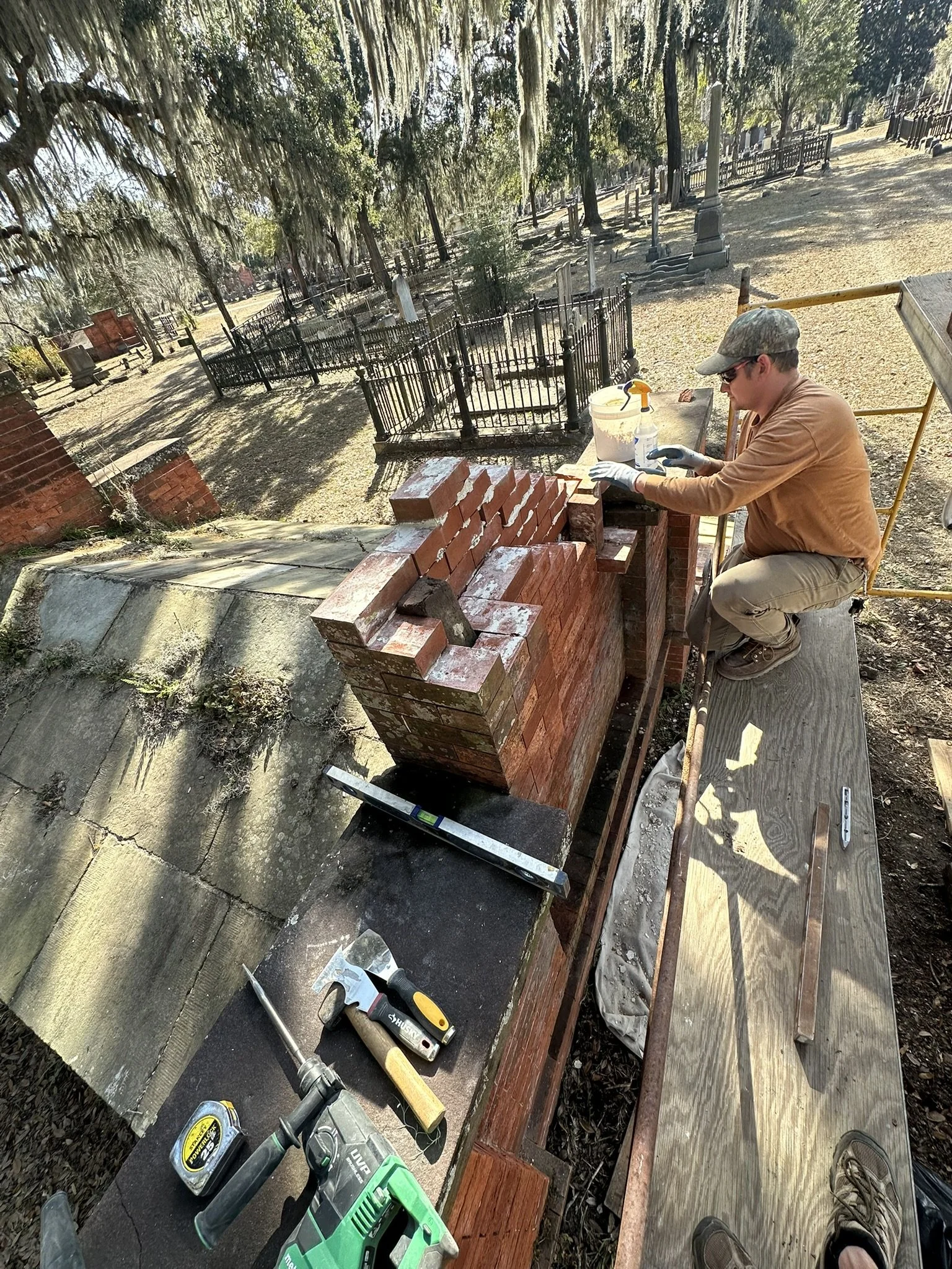 A person working on a brick wall outdoors, surrounded by gravestones and trees in a cemetery. The person is wearing gloves, sunglasses, a cap, and tan clothes, and is using tools such as a hammer and power drill for construction.