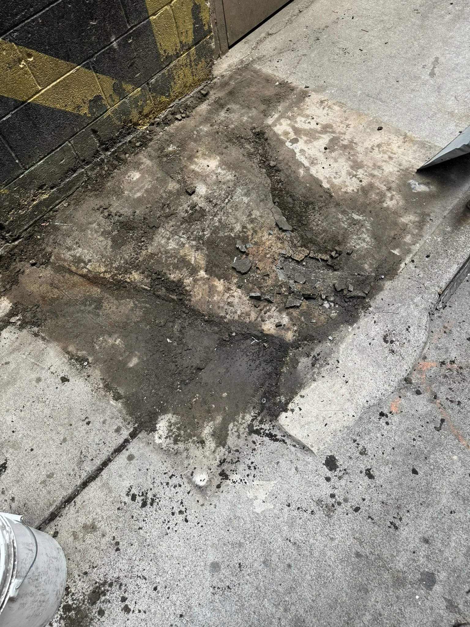 Part of an indoor concrete floor with freshly cut and excavated square from the asphalt, surrounded by scattered dirt and debris, with a wall partially painted with black and yellow checker pattern.