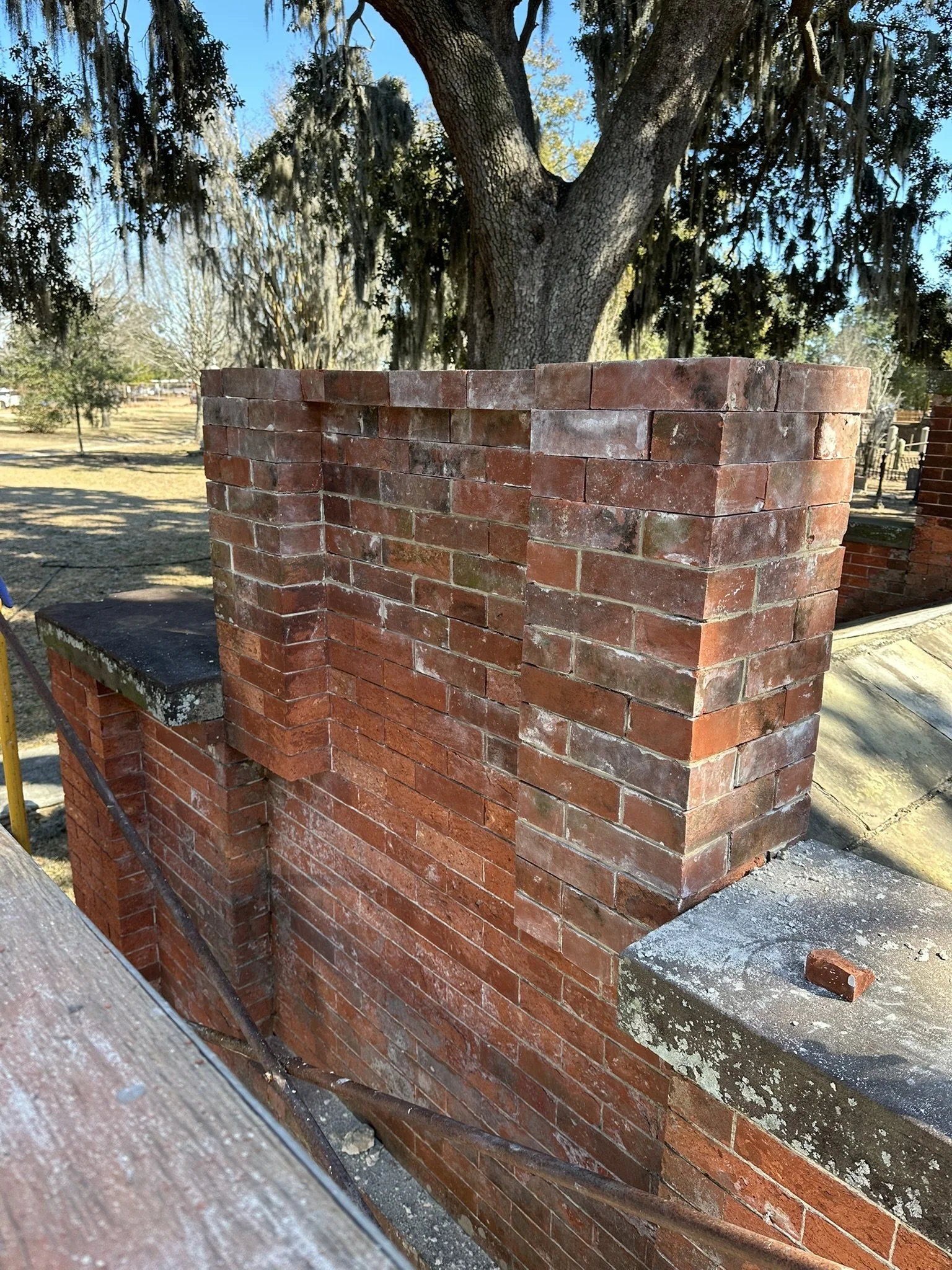 Red brick chimney under construction with a large tree in the background and a clear blue sky.