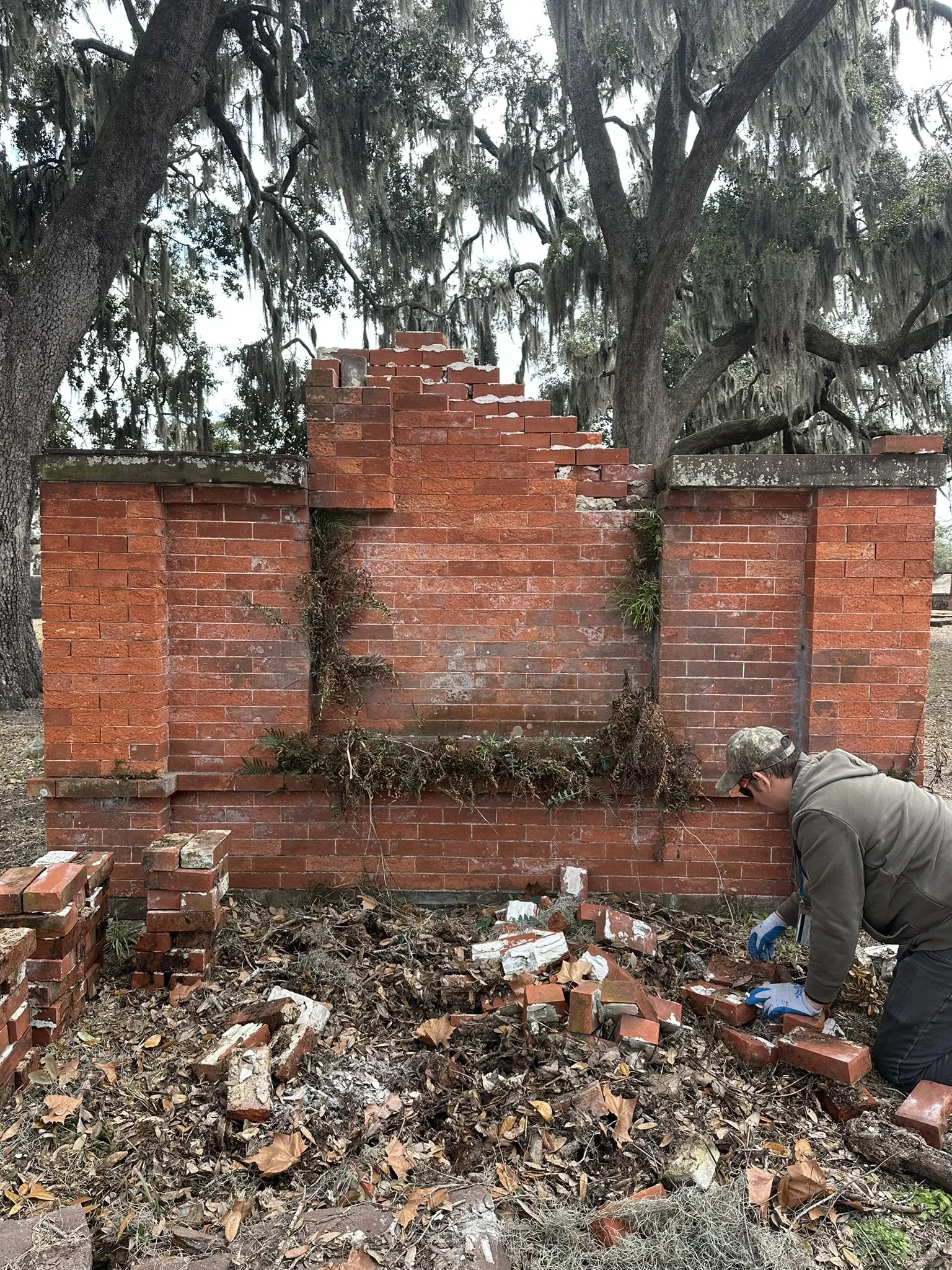 Person wearing gloves and a hoodie kneeling down, working on a pile of broken bricks and debris in front of a partially damaged brick wall with large trees in the background.
