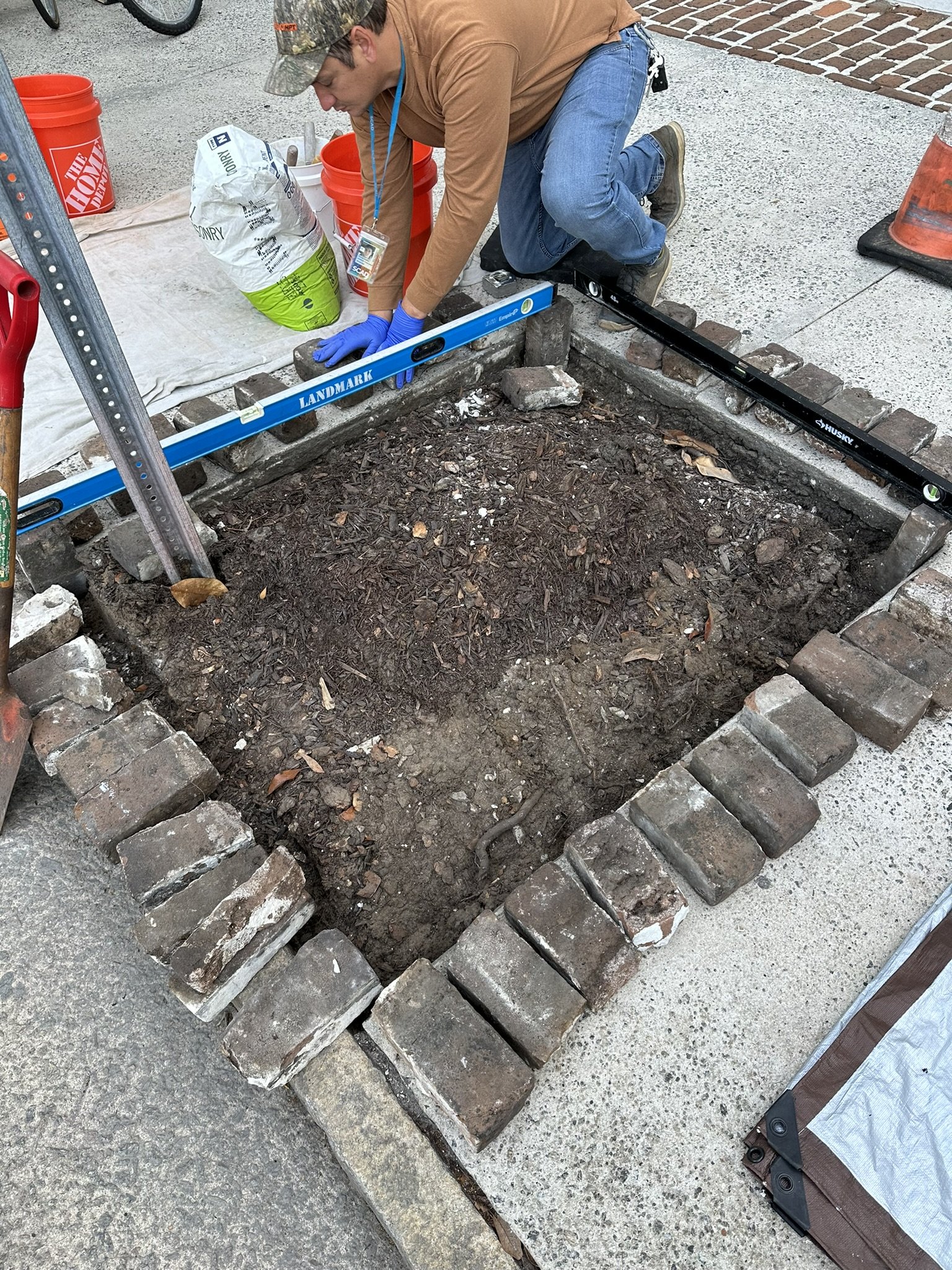 A person kneeling on the ground working on a brick-lined garden bed or pathway, with tools and construction materials around.
