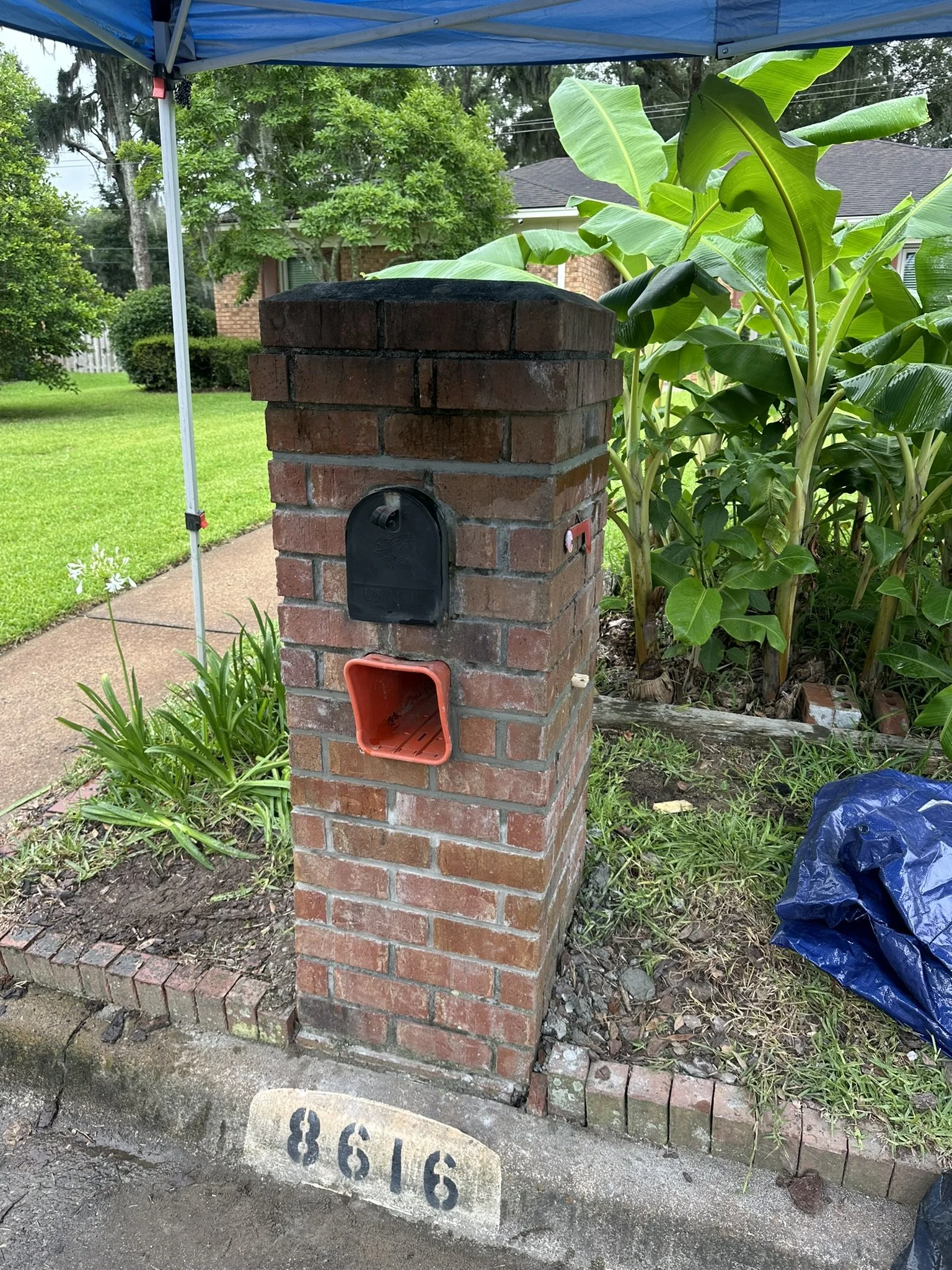 A brick mailbox with a black metal door and a red plastic container below it, situated on a sidewalk next to a garden bed with green plants, and a house in the background with trees and a blue canopy above.