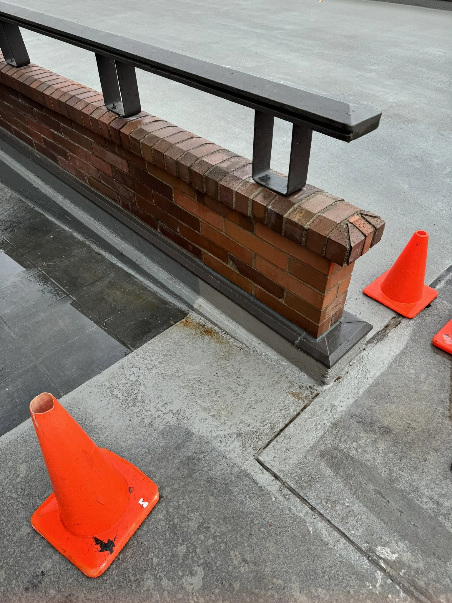 Orange traffic cone with black scuff marks in front of a brick and metal barrier on a sidewalk.