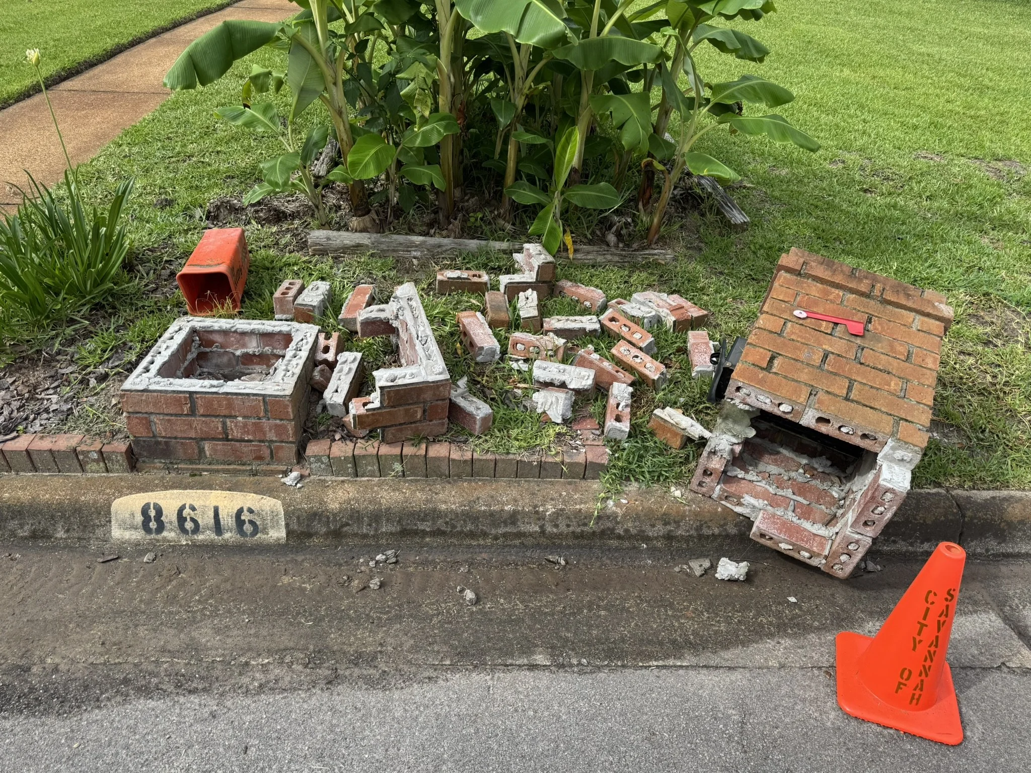 A sidewalk and street with a black curb separating them. There is a construction or repair zone with a small brick wall partially built, scattered bricks, an orange safety cone with a warning label, and a horizontal brick slab on the ground. In the background, there is a grassy area with a shrub and a row of green plants, including banana plants.