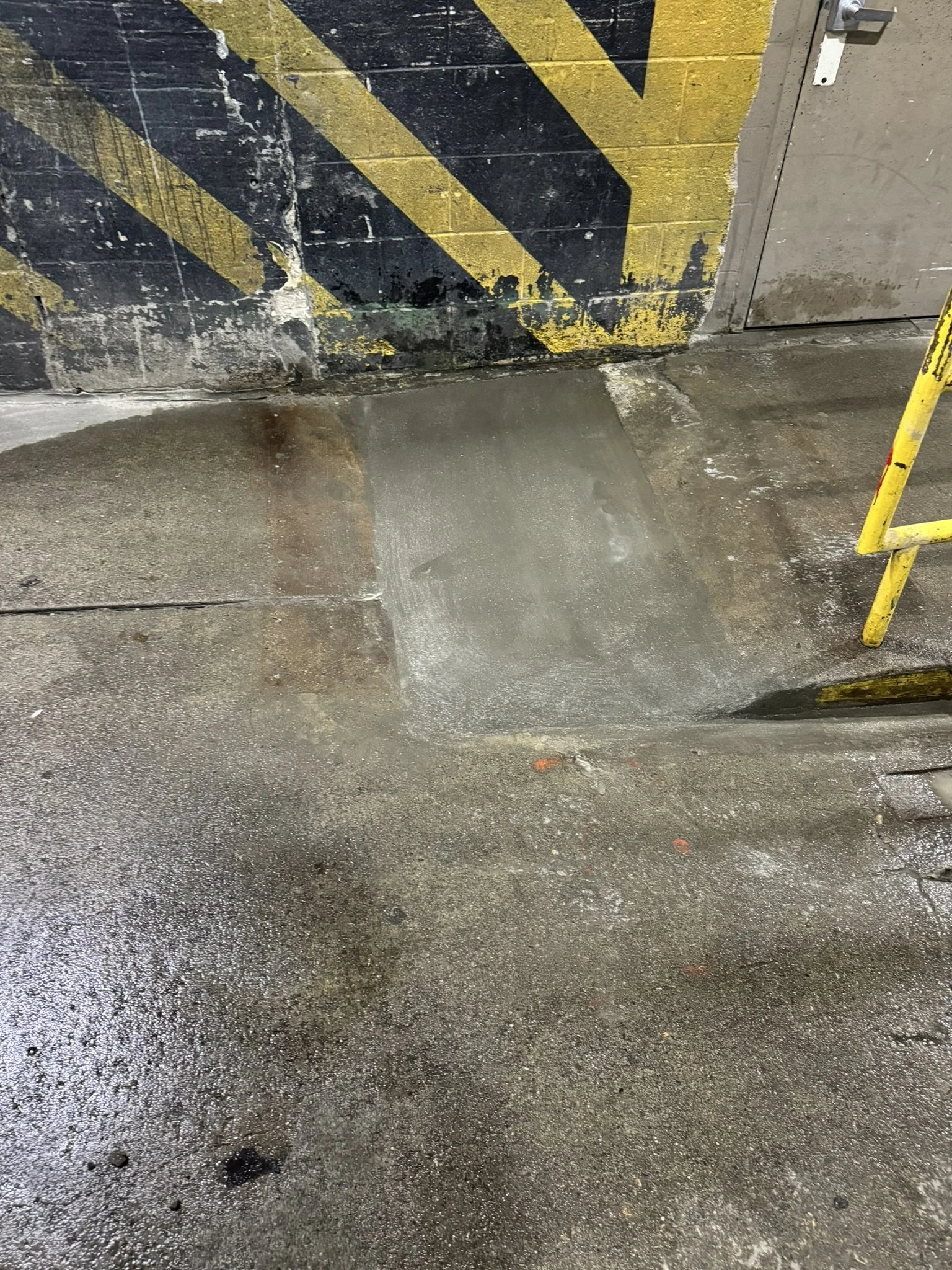 Wet concrete floor in an industrial or parking garage setting with a black and yellow striped hazard marker on the wall and a yellow metal barrier on the right.
