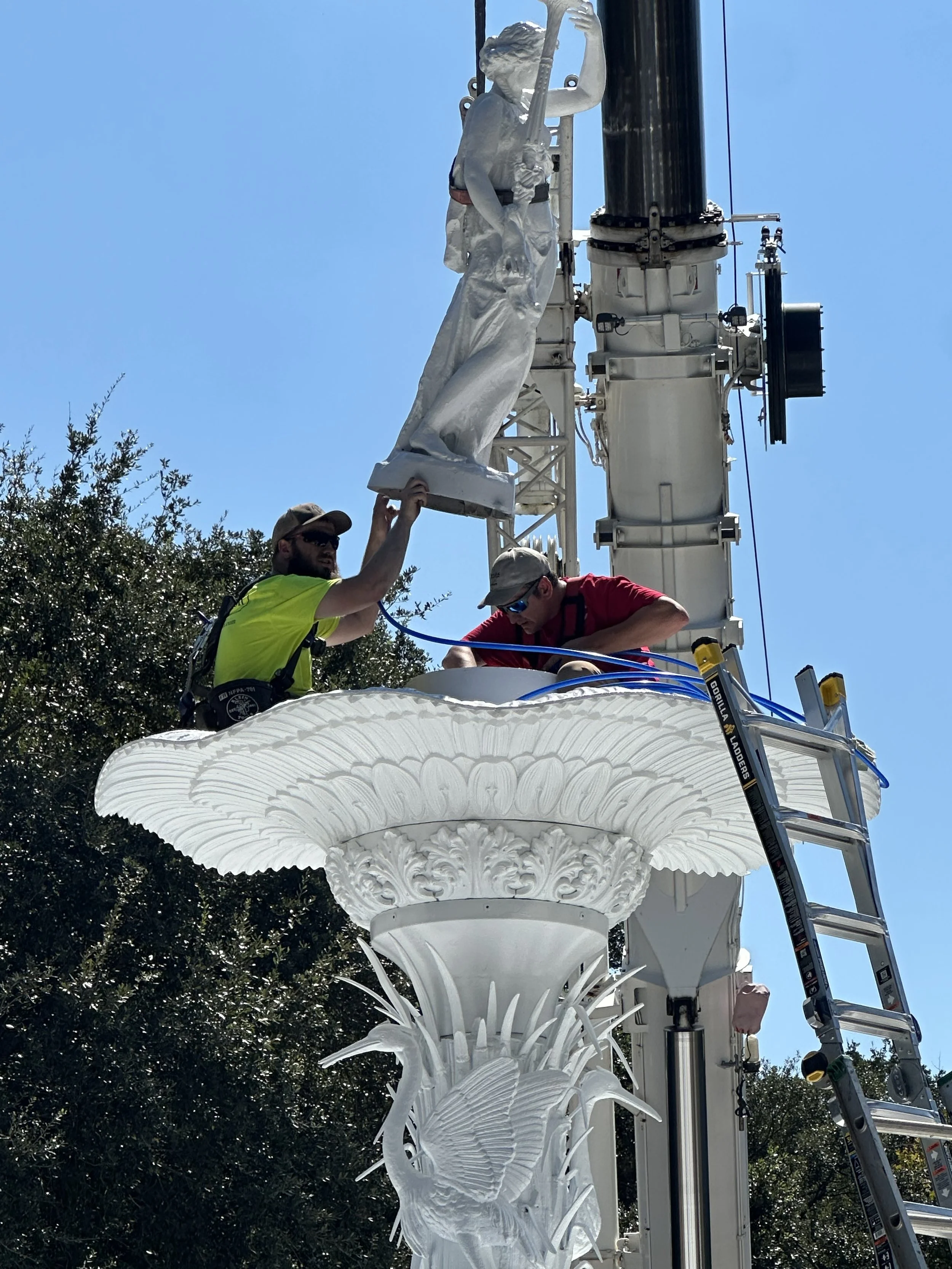 Two workers installing or maintaining a statue of a woman with a musical instrument on a decorative streetlight pole, with greenery and a blue sky in the background.