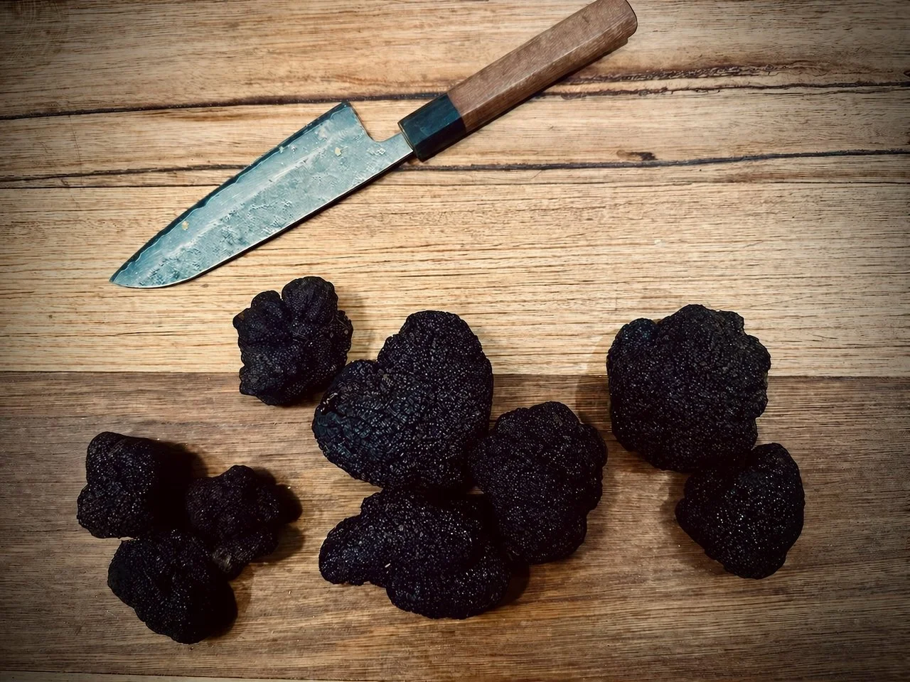 A knife with a wooden handle and metal blade lying on a wooden surface with several black truffles arranged below it.