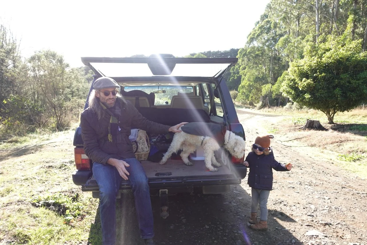 A man with long hair, wearing sunglasses, a cap, and a dark jacket, is sitting on the bumper of Range Rover Classic car, with a Lagotto Ramagnolo dog standing on the car's trunk. On a dirt path, with trees and greenery in the background.