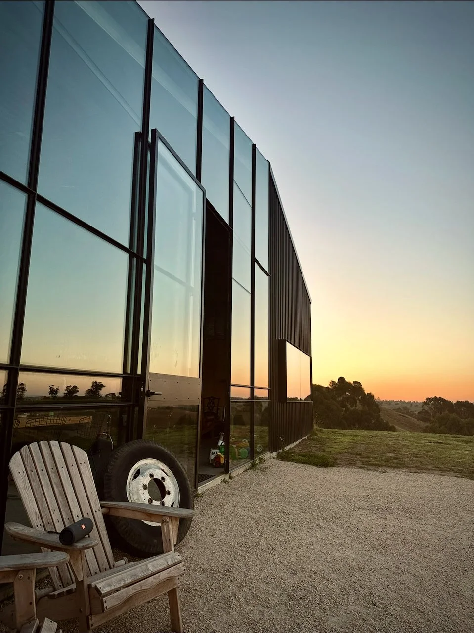Modern building with large glass windows reflecting sunset, wooden chairs, and an old tire outside, overlooking a grassy landscape at sunset.