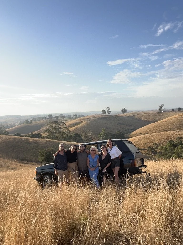 Group of six women standing in a grassy field with hills and trees in the background, next to a Range Rover Classic under a perfect early evening sky.