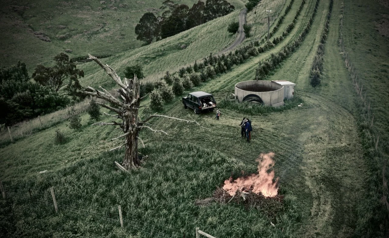 Aerial view of a rural landscape, a Truffiere with a dead tree, a fire burning in a pile of wood, and a Range Rover Classic vehicle parked near the tree. Two people are walking on a grassy path.