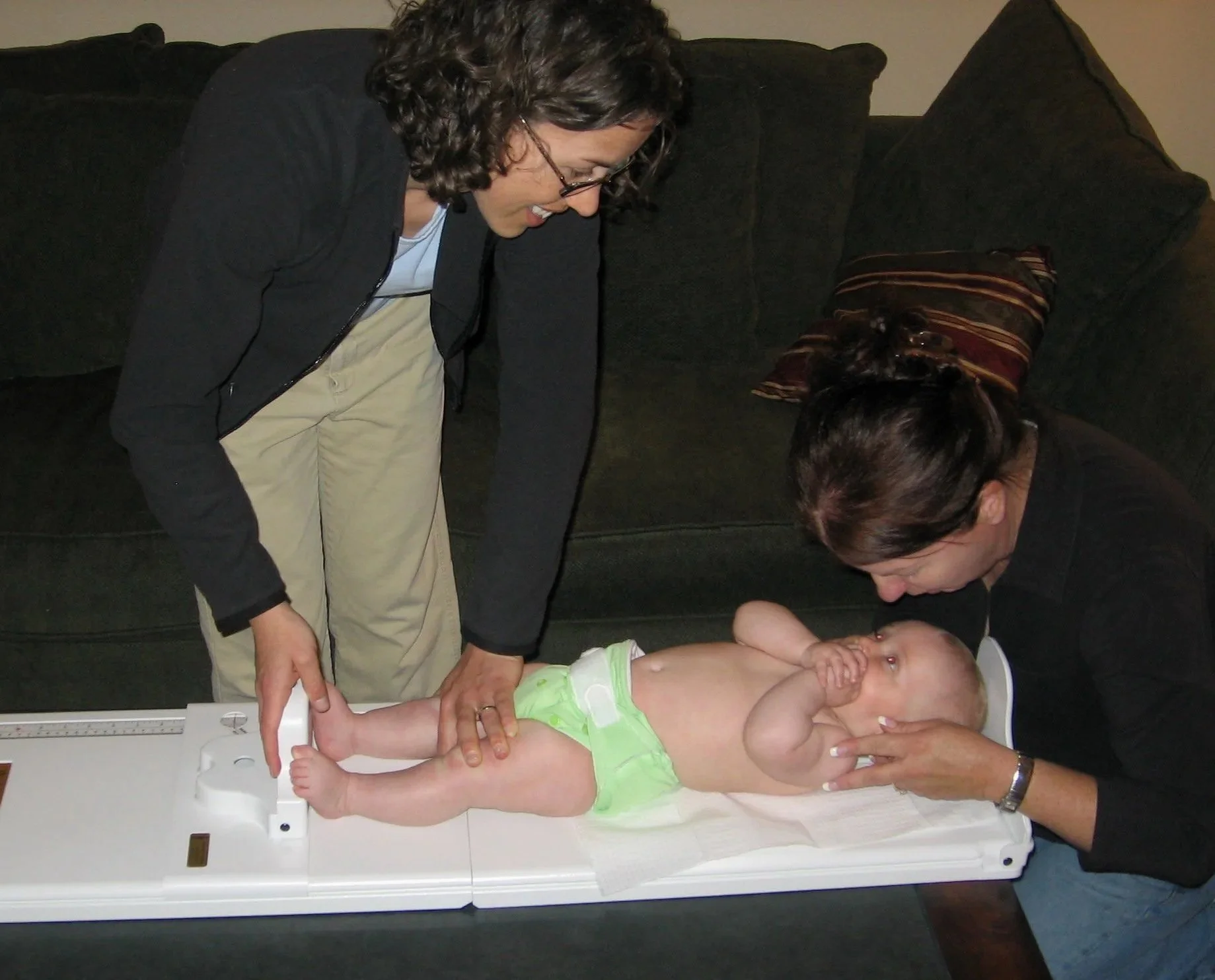 A woman and a man are measuring the length of a baby lying on a scale in a medical setting. The woman is standing and looking down at the baby, while the man is leaning over, holding the baby's leg, and looking at the baby. The baby is wearing a light green outfit and is resting on a white medical scale in a room with a dark couch and a striped pillow in the background.