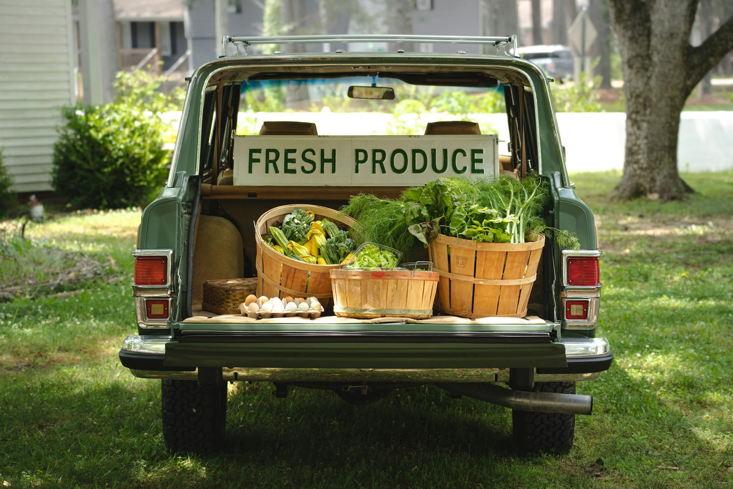 The back of a green vintage vehicle filled with farm fresh produce such as eggs, leafy greens, zucchinis, and other vegetables, with a sign reading 'FRESH PRODUCE' displayed inside.