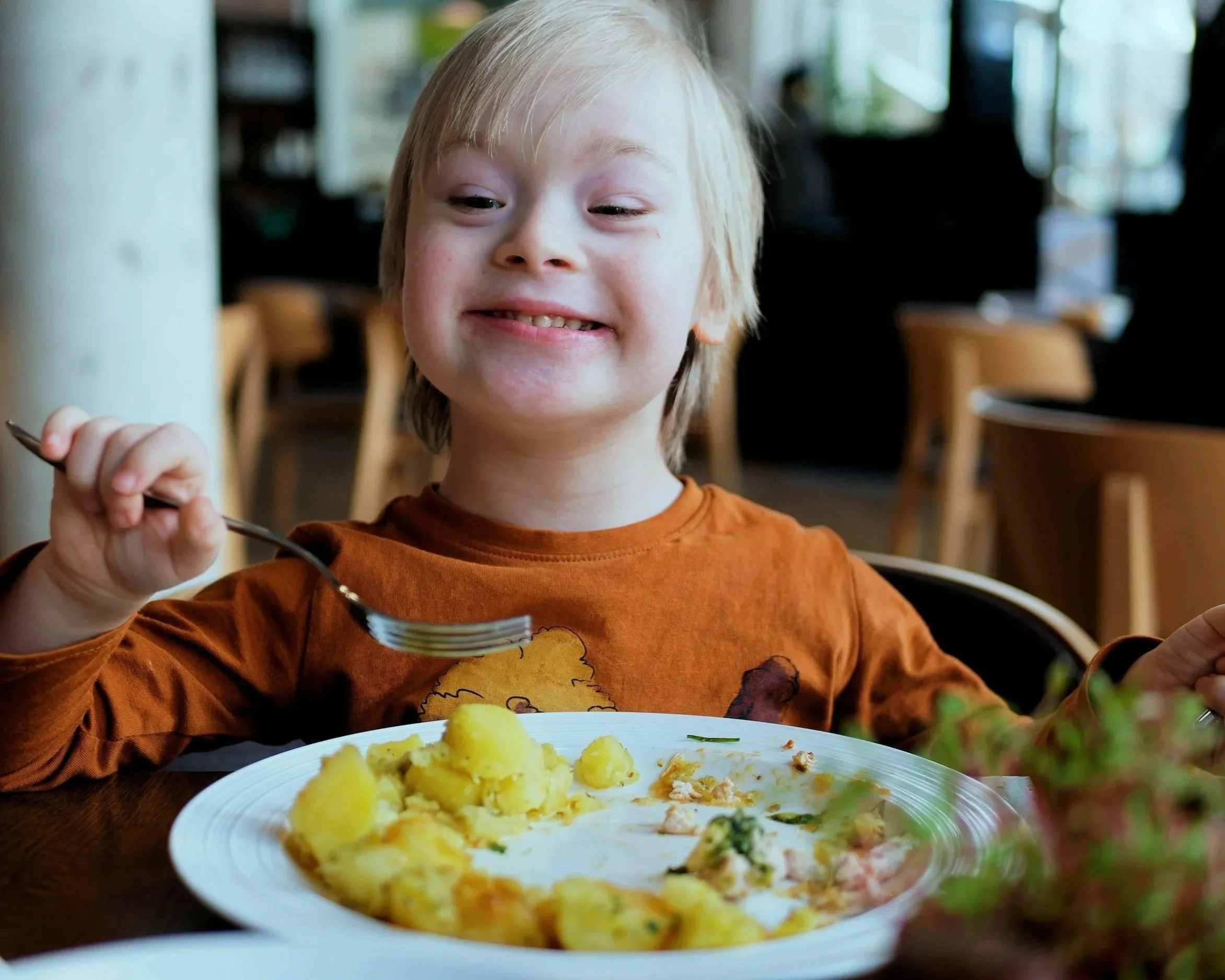 Young boy with blonde hair smiling while holding a fork at a table with a plate of mashed potatoes and vegetables in a restaurant or cafe setting.