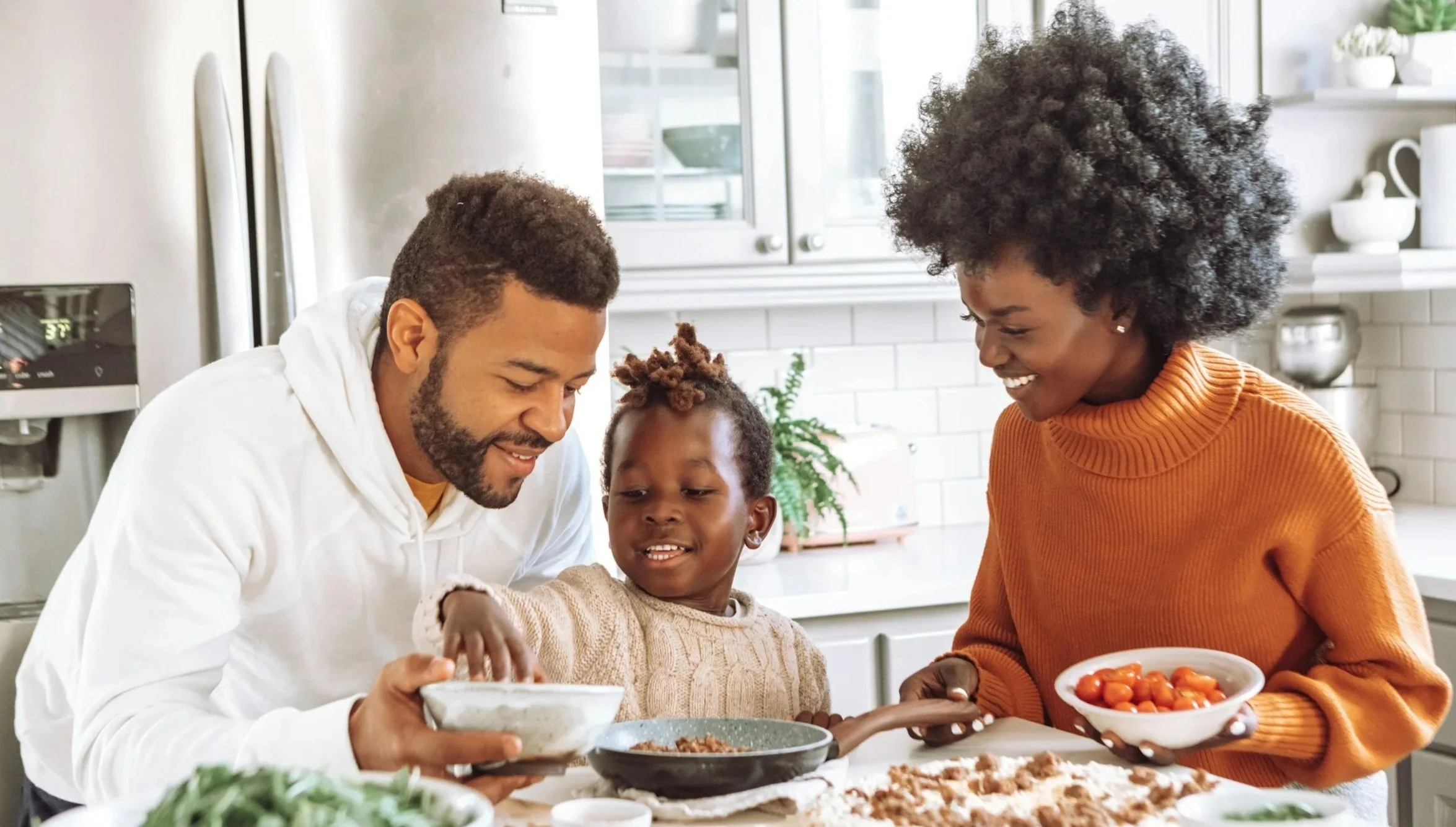 Family preparing and sharing a meal in a modern kitchen, with a man, woman, and young girl smiling and serving food.