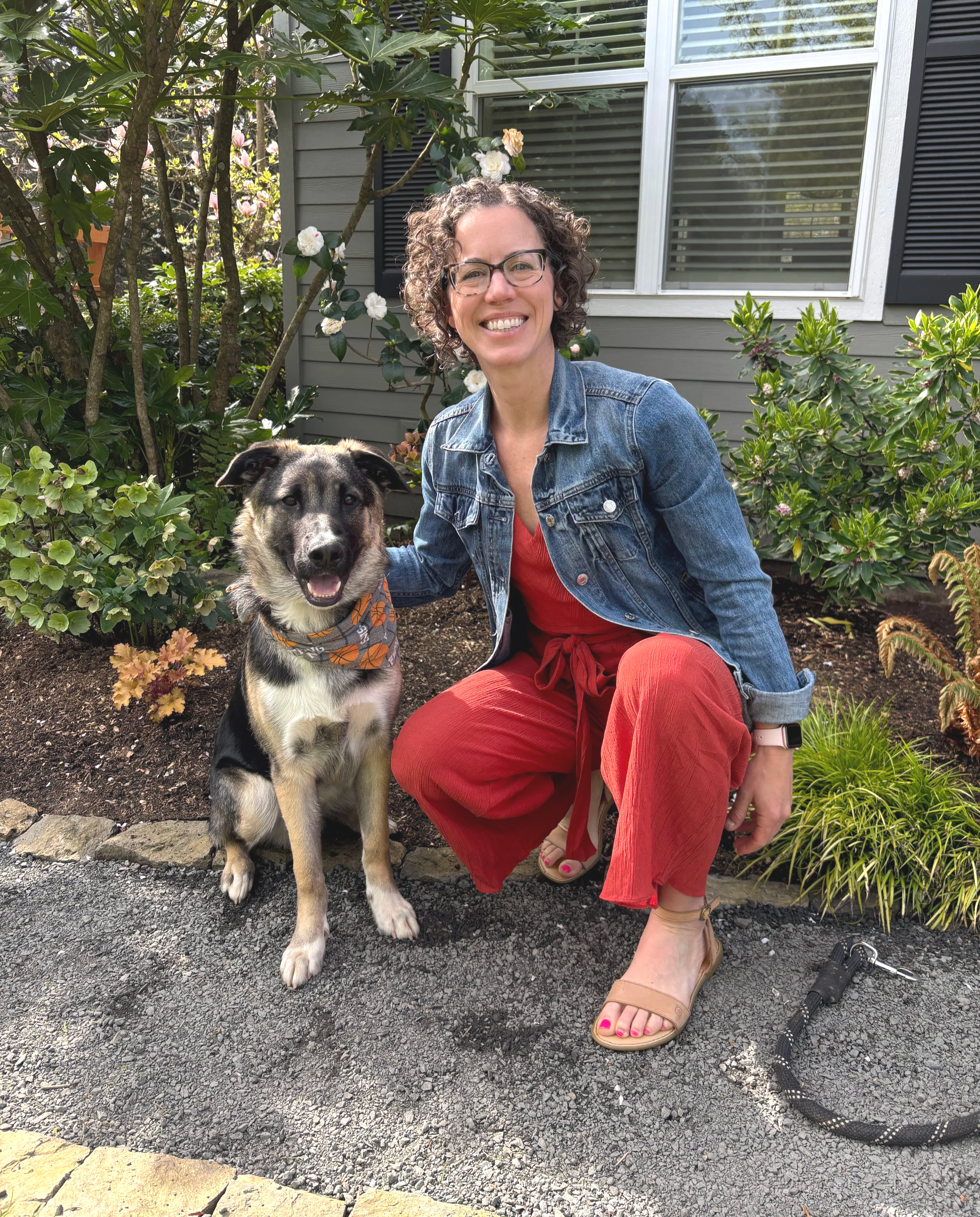 A woman with curly hair, glasses, wearing a denim jacket and red pants, smiling and kneeling next to a black-and-white shepherd dog with a bandana, in a garden in front of a gray house with a window, surrounded by green plants and bushes.