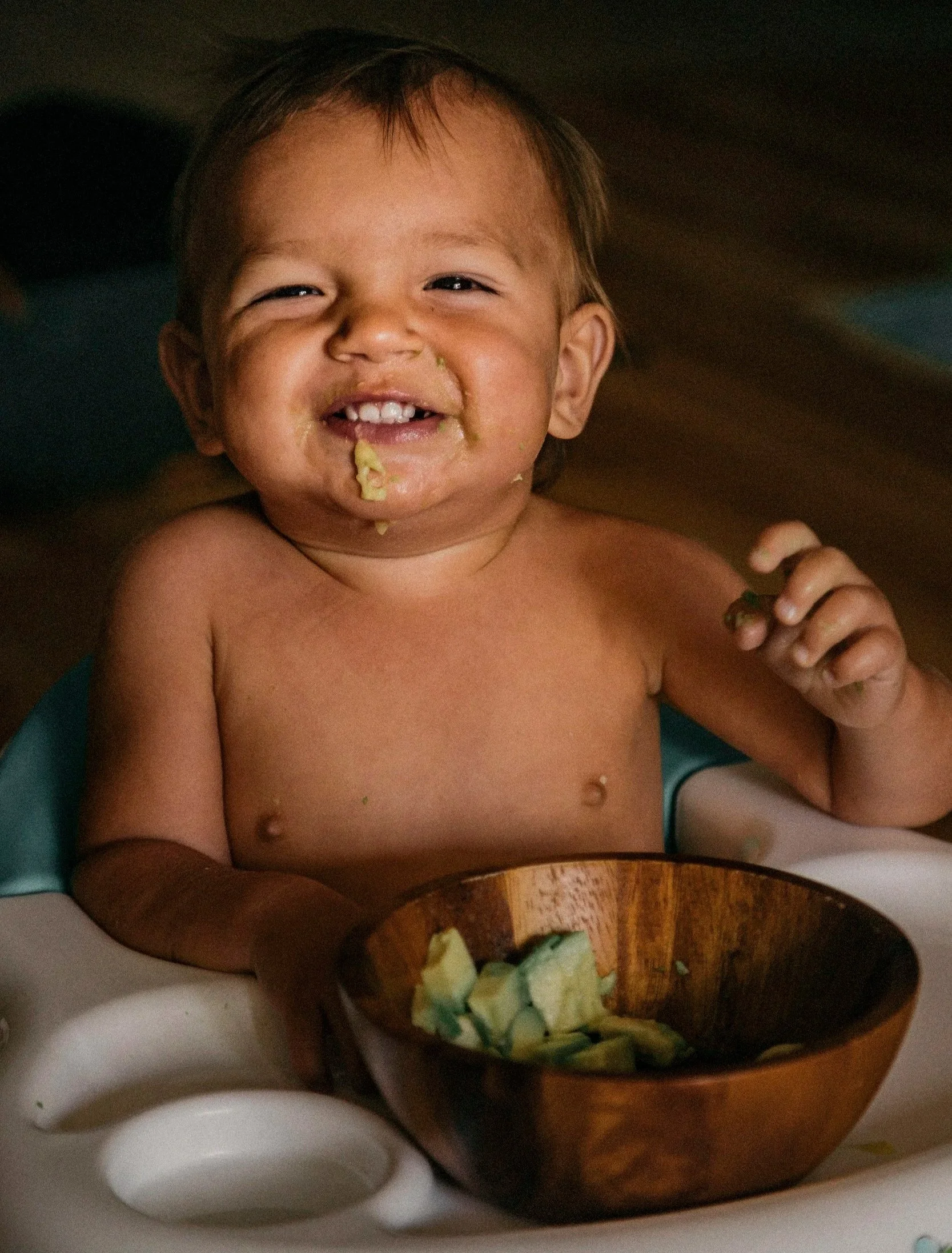 Young boy with short brown hair smiling with food around his mouth, sitting at a table with a wooden bowl of chopped vegetables in front of him.