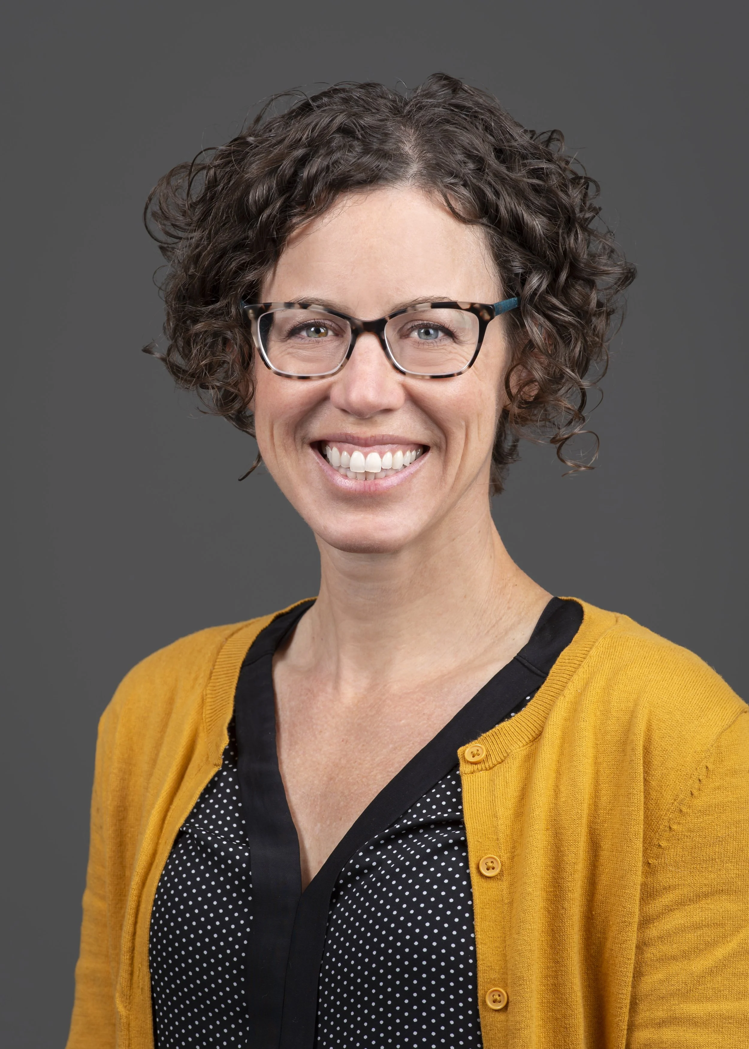 A woman with short curly brown hair, wearing glasses with tortoiseshell frames, a black polka dot blouse, and a mustard yellow cardigan, smiling against a plain gray background.
