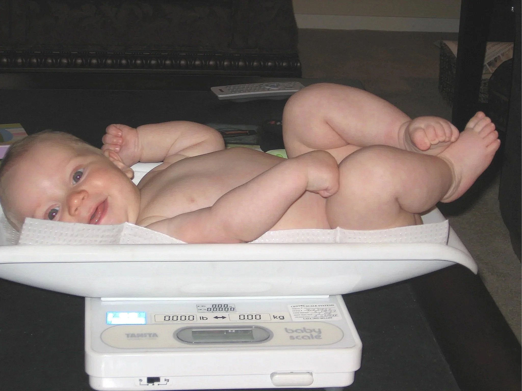 A digital baby scale with a newborn baby lying on it, the baby is looking at the camera with one hand near the face and the other on the body.