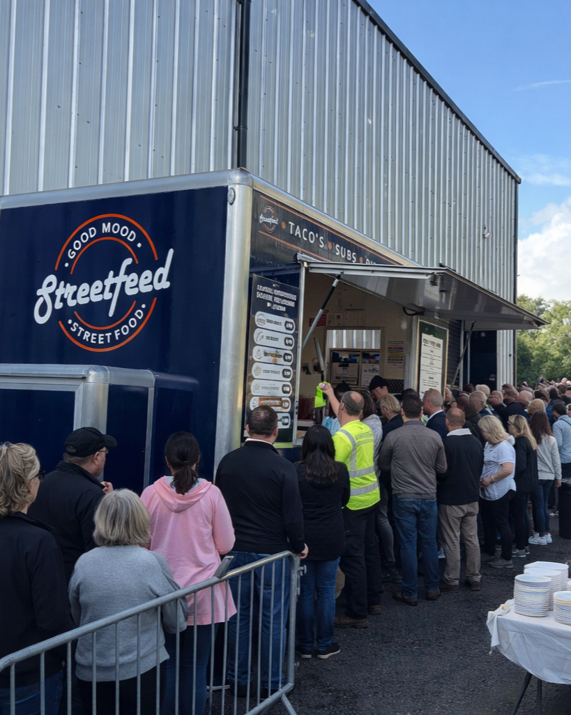 A long line of people waiting outside a food truck that has the words 'Streetfeed' and 'Good Mood Street Food' on its side. The truck offers tacos, subs, and other street food, with a menu displayed on the side. The scene is outdoors and sunny, with a large metal building in the background.