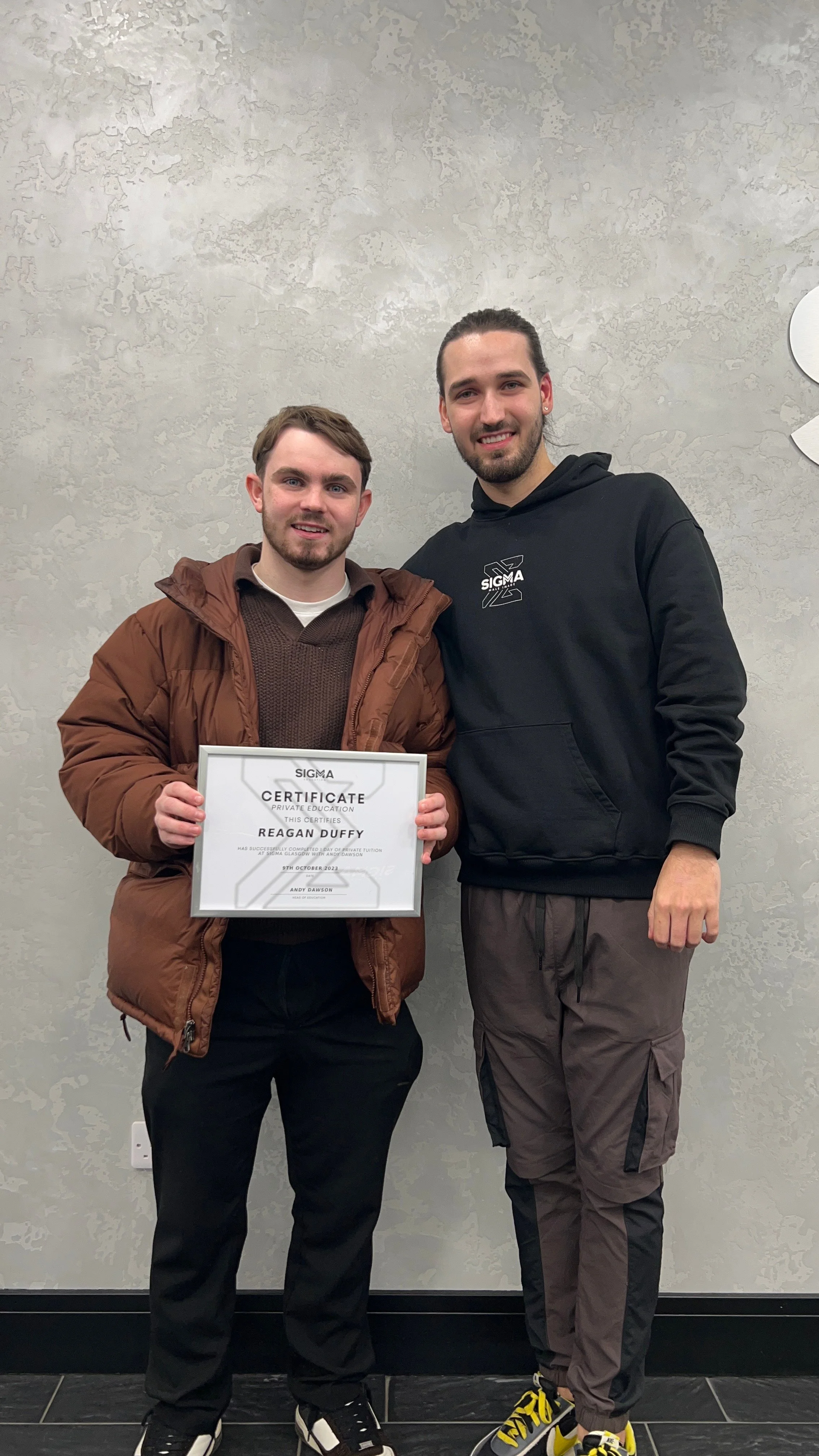 Two young men standing side by side on a gray wall background, one holding a certificate. The man on the left wears a brown jacket and black pants, while the man on the right wears a black hoodie and brown pants with black and yellow shoes. They are 