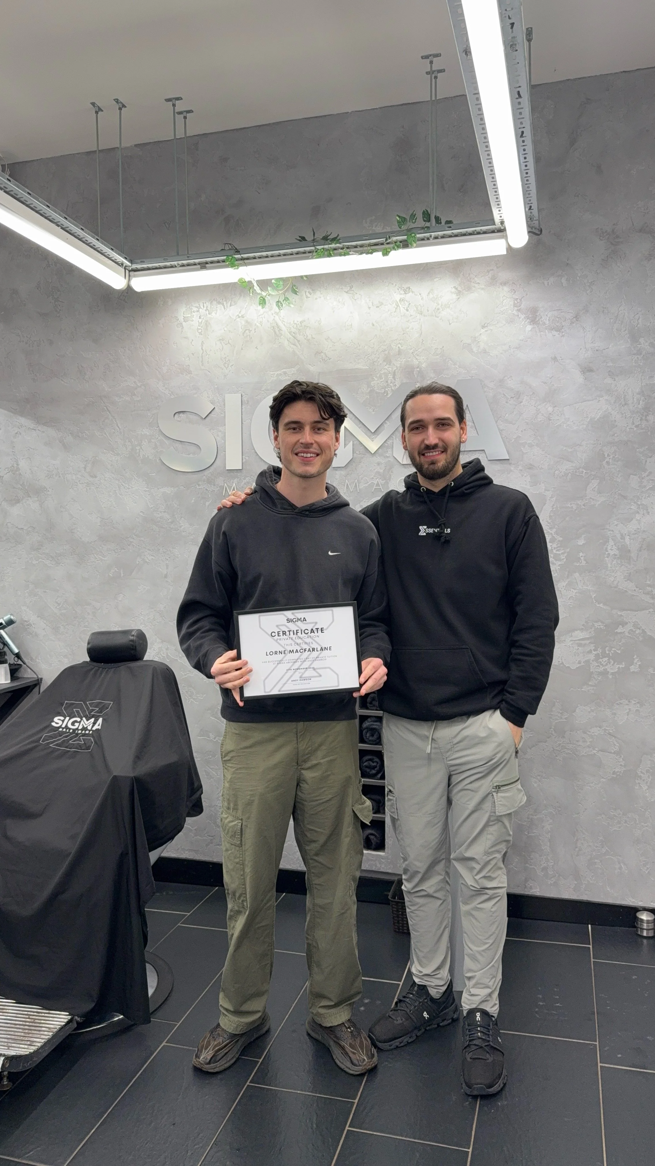 Two young men standing inside a modern office, one holding a certificate, smiling at the camera. Behind them is a gray wall with the Sigma logo, and a desk with a covered chair is visible to the left.