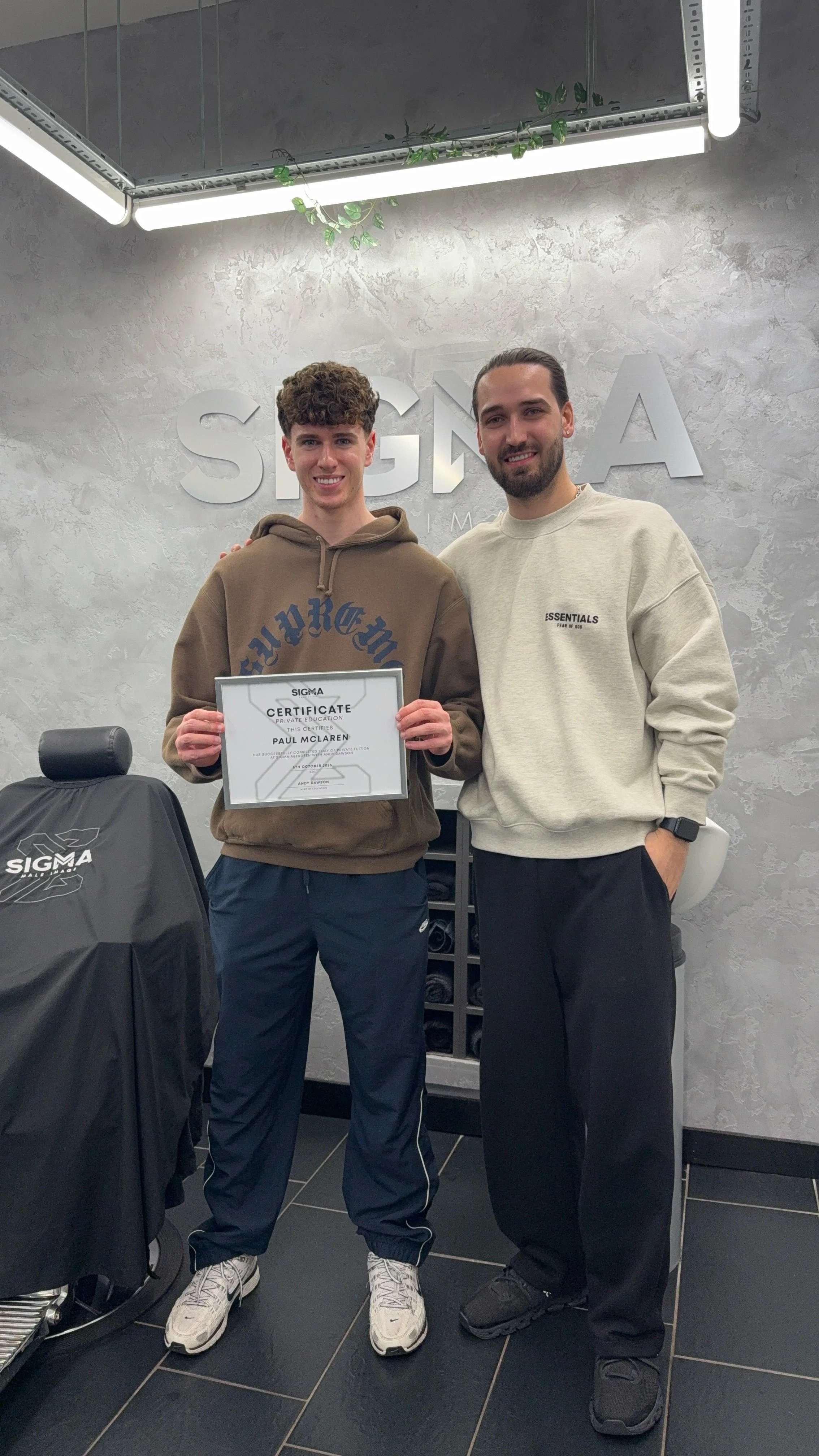 A young man in a brown hoodie holding a certificate standing next to an older man in a beige sweatshirt. They are inside a room with a gray textured wall that has 'SIGMA' written on it. The young man is smiling and the older man has his arm around hi