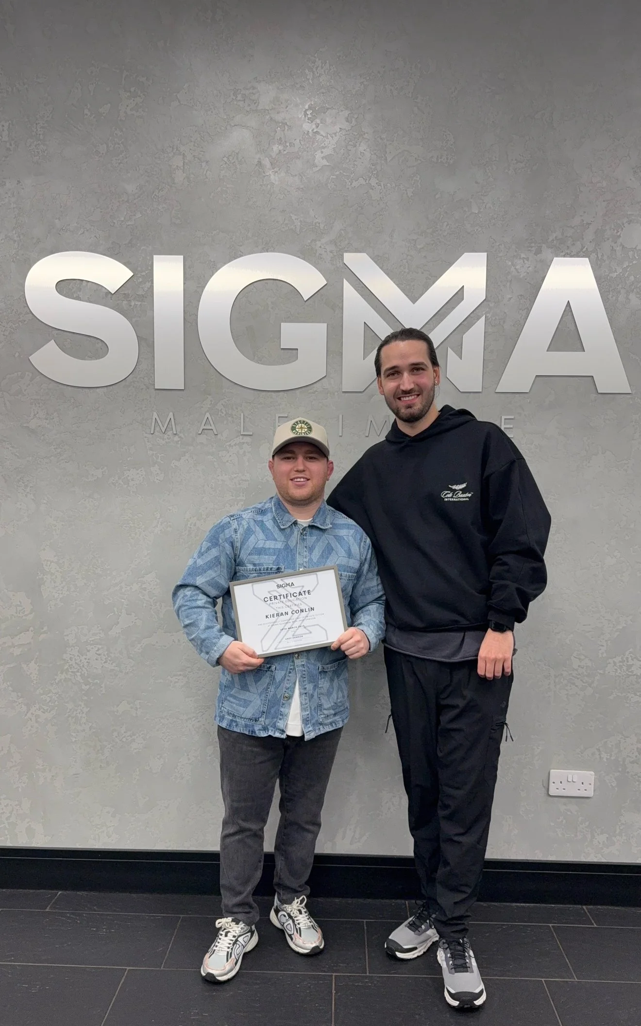 Two young men standing side by side in front of a textured wall with large letters spelling 'SIGMA' and 'MAL' beneath, one holding a certificate of achievement.