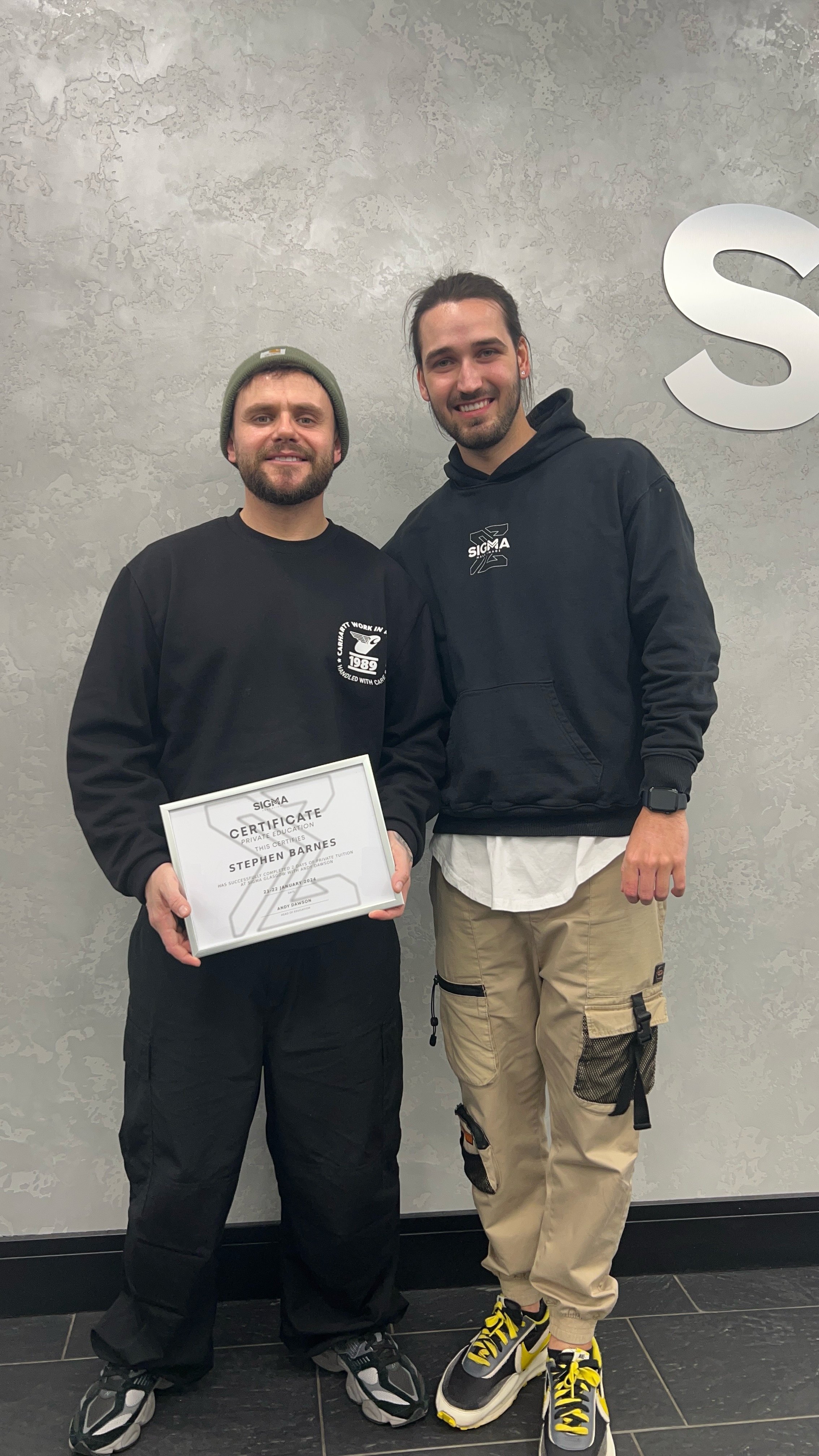 Two men standing indoors, one holding a certificate. The man on the left has a beard, is wearing a beanie and black sweatshirt. The man on the right has long hair, is wearing a black hoodie, khaki cargo pants and sneakers.