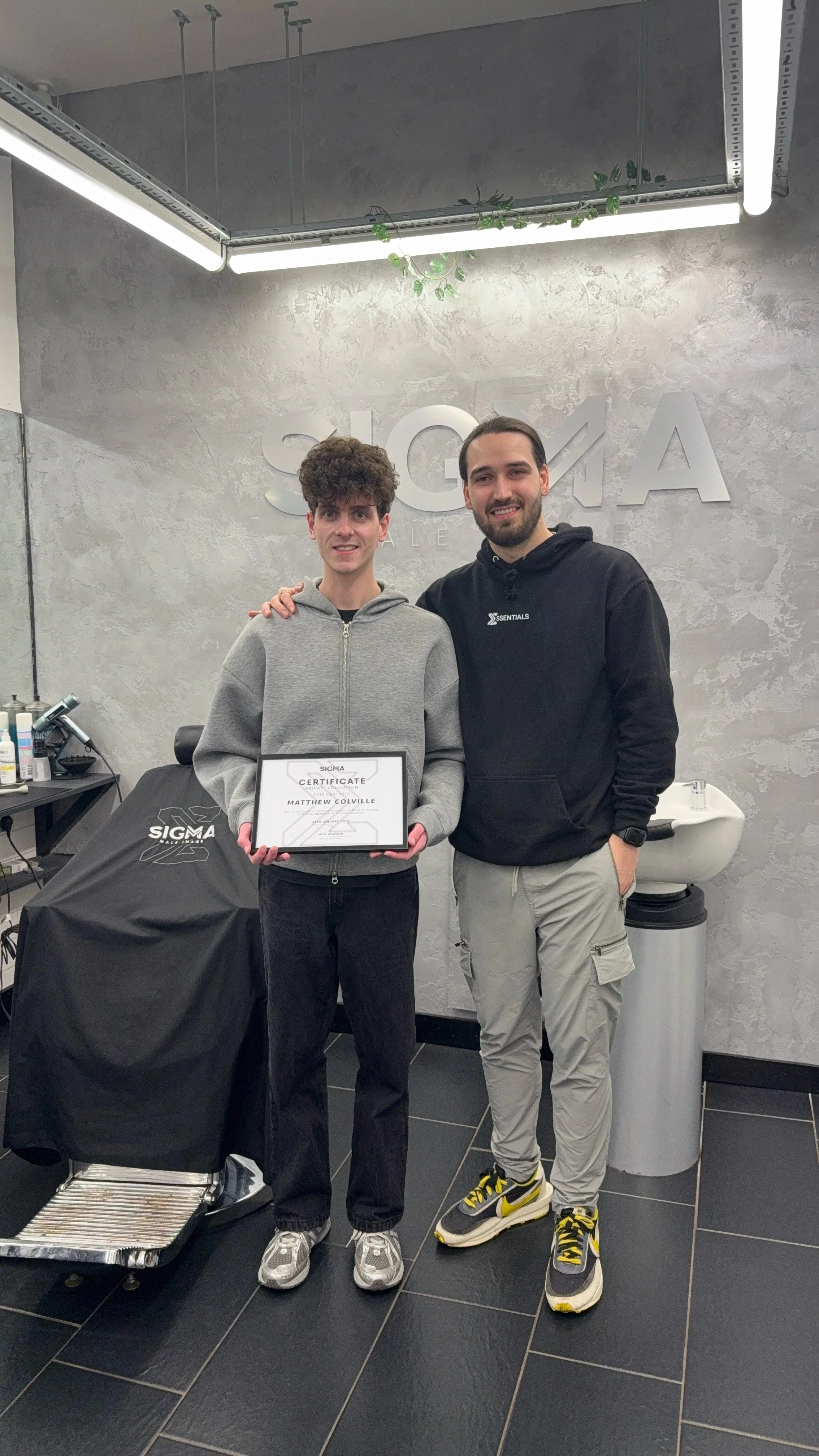 Two young men standing in a hair salon, smiling for a photo. The person on the left holds a certificate, and the person on the right has his arm around the other. The salon has a modern, industrial design with the Sigma logo on the wall behind them.