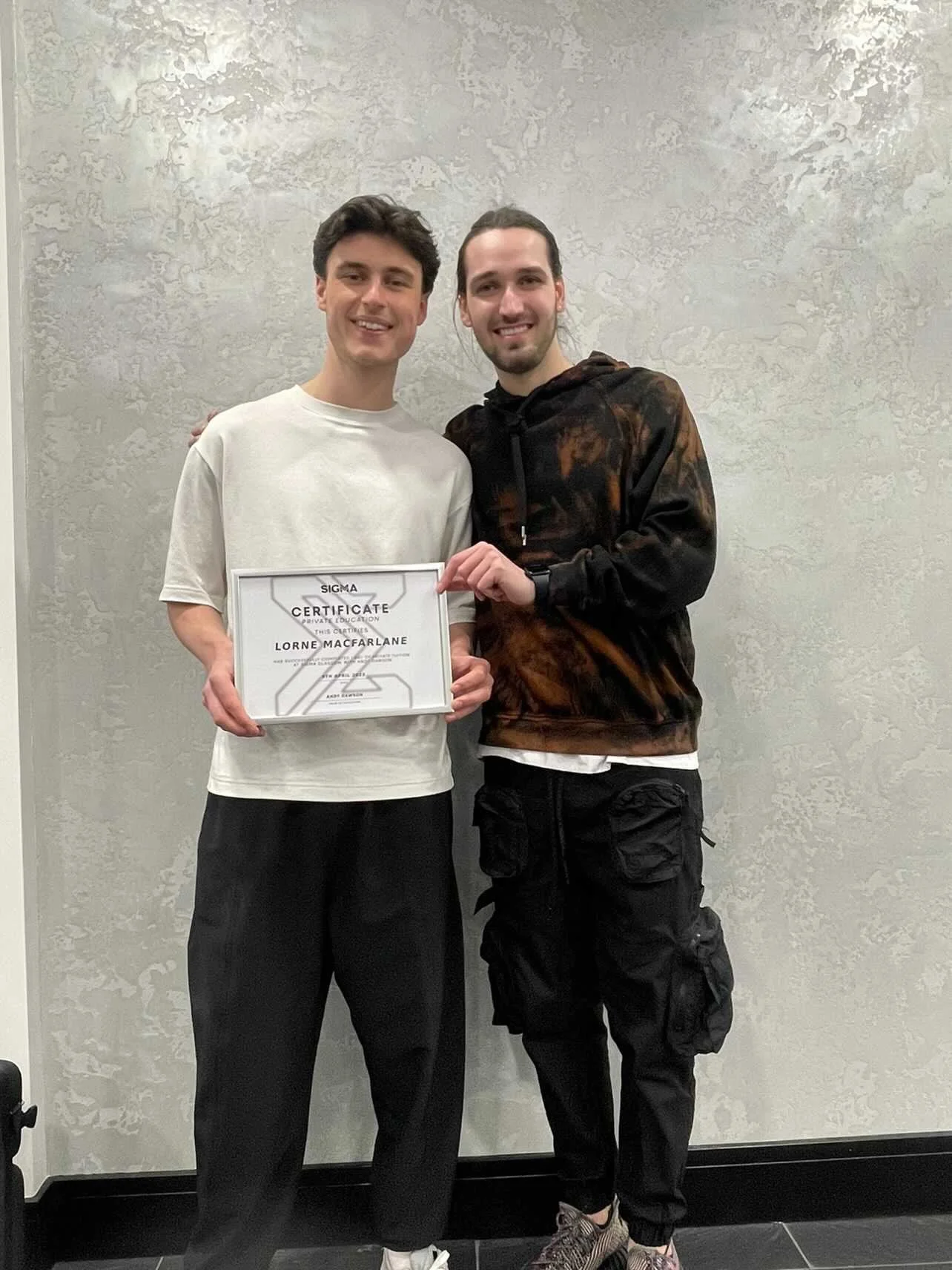 Two young men smiling, standing side by side against a textured gray wall, with the one on the left holding a certificate for Lorne MacFarlane from Sigma.