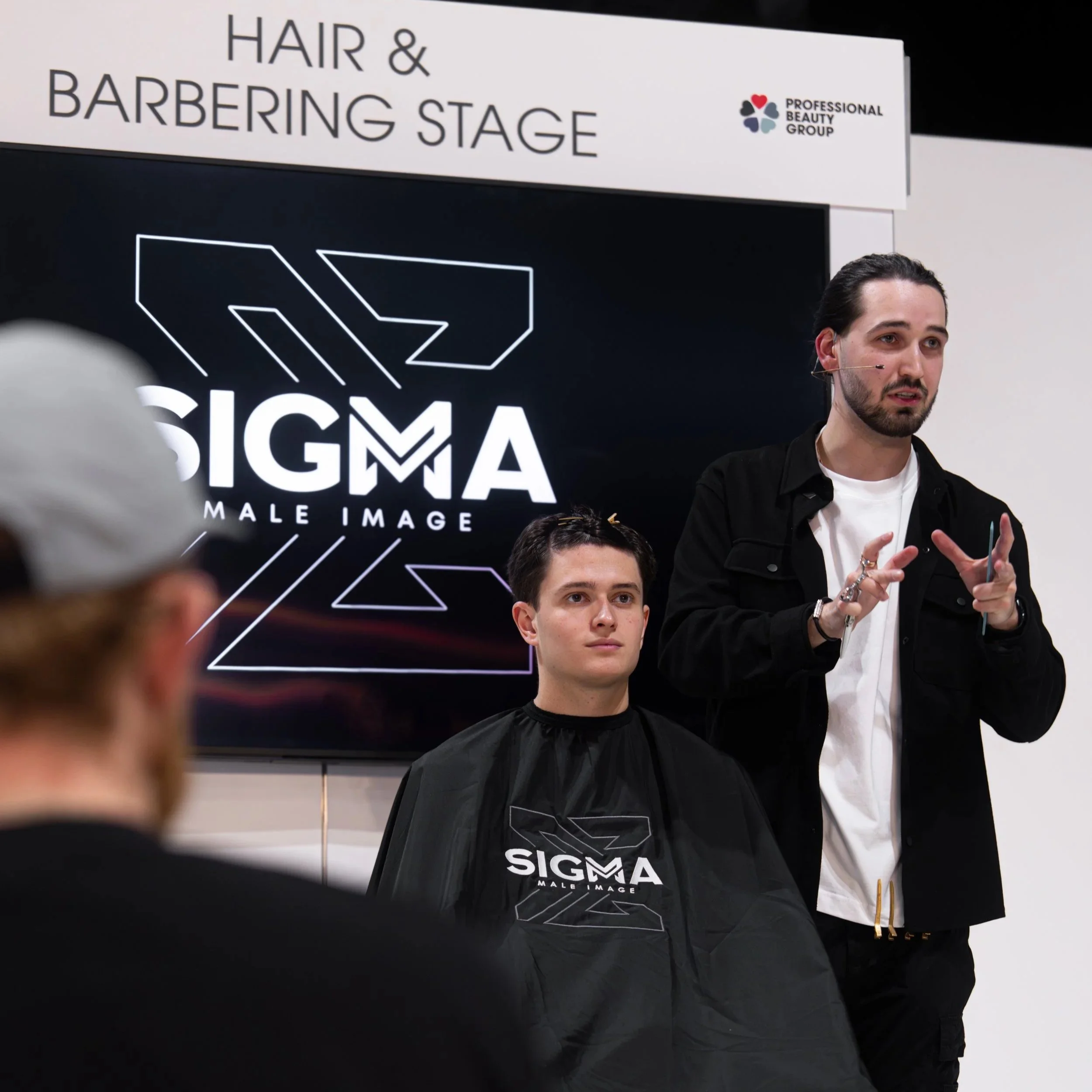 A male hairstylist giving a presentation at a workshop labeled 'Hair & Barbering Stage' with a large Sigma Male Image logo on the screen behind him. A young man sits in a barber's cape with the same logo, listening attentively.